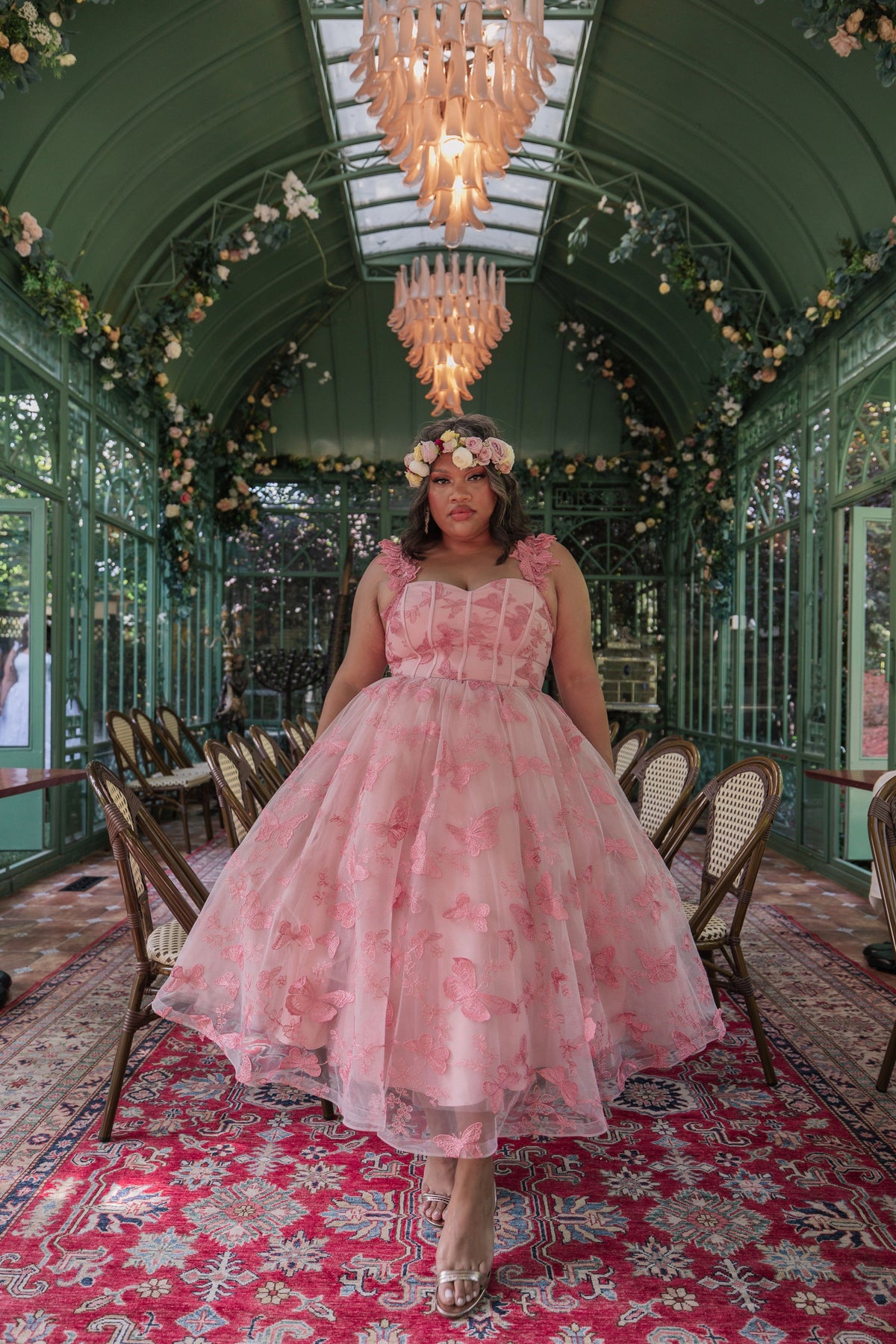 The image shows a woman in a pink dress standing on a red carpet in a room with chairs, a chandelier, and a glass wall in the background.