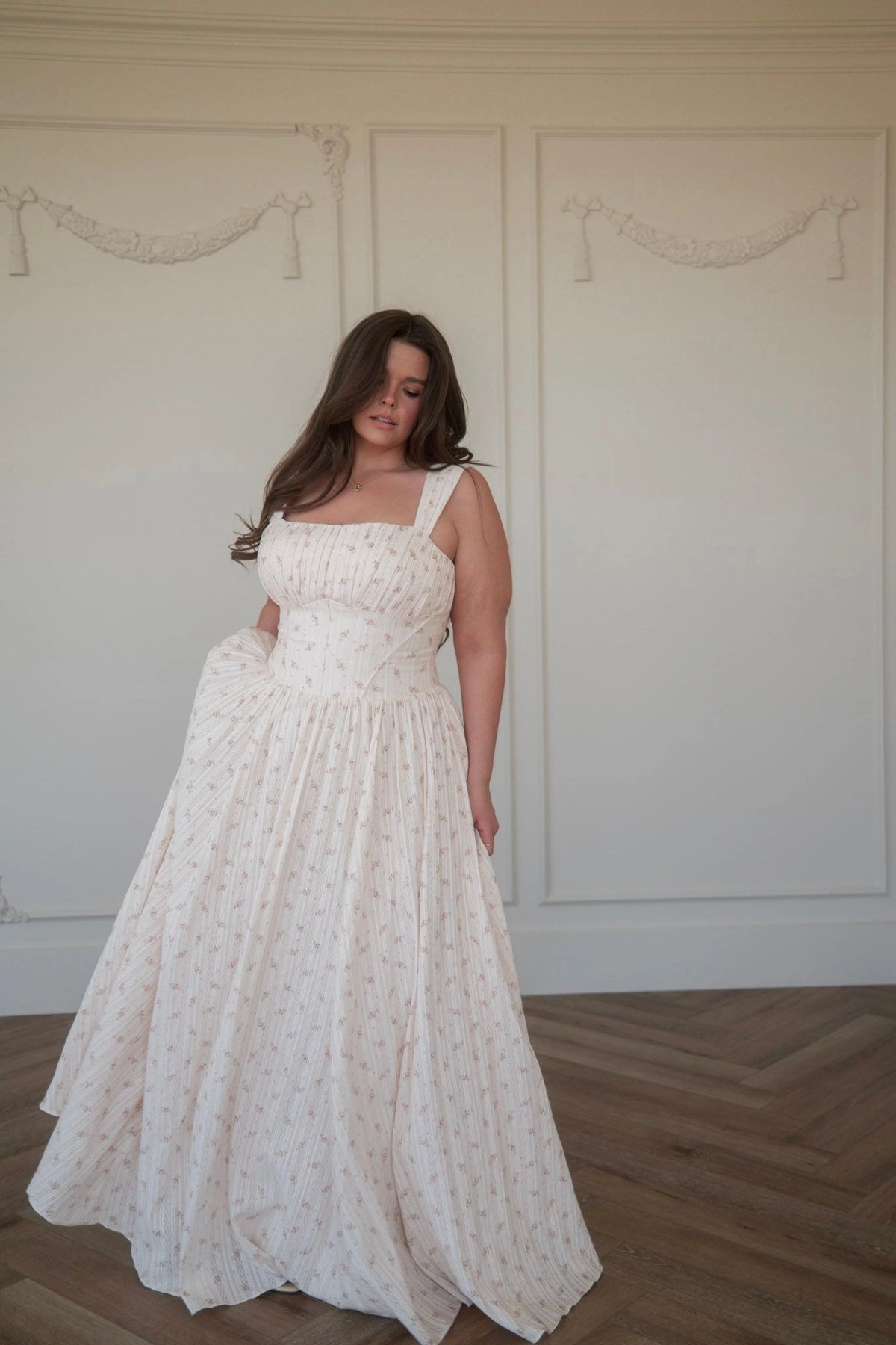 Woman in white sleeveless gown with square neckline and subtle pattern stands in front of white wall, looking down with right hand on hip.