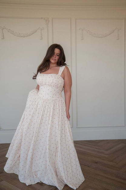 Woman in white sleeveless gown with square neckline and subtle pattern stands in front of white wall, looking down with right hand on hip.