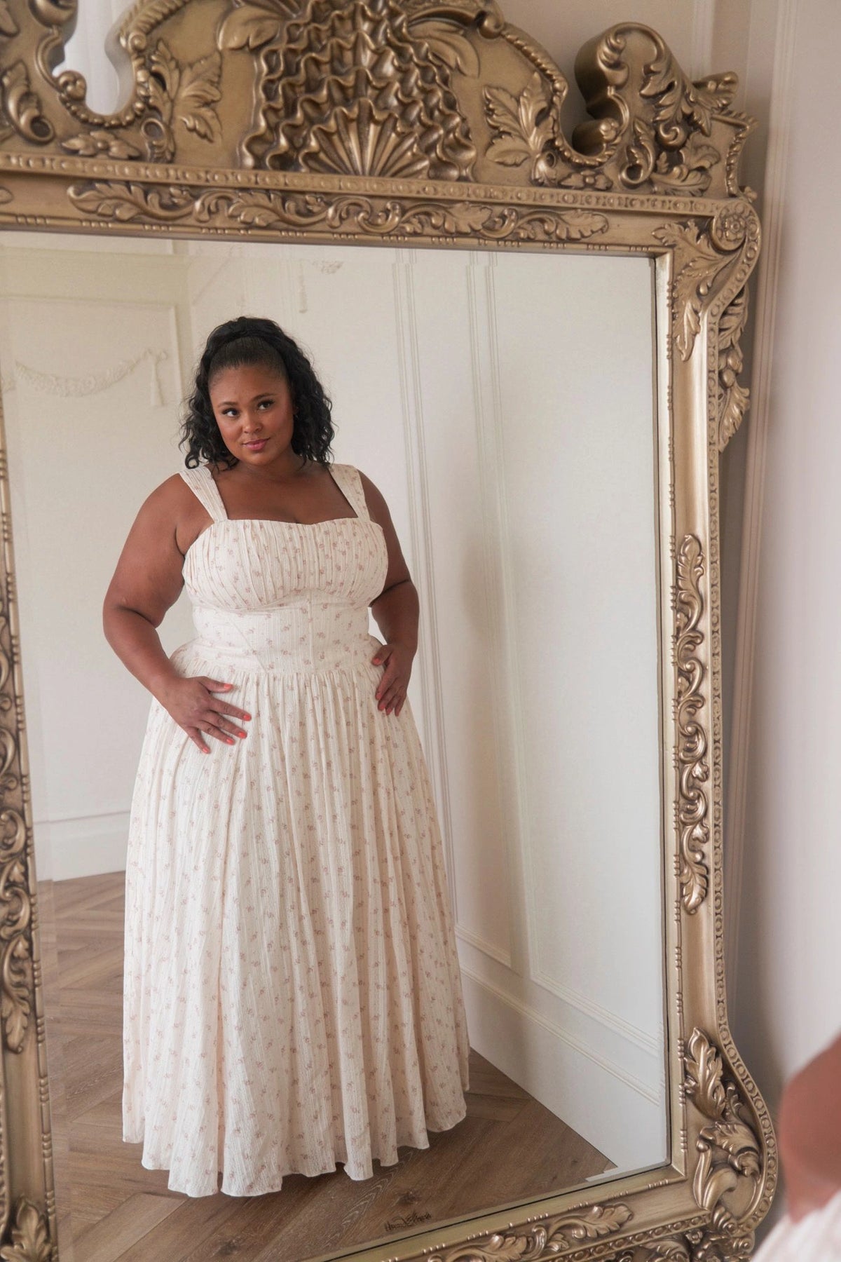 The image shows a woman in a white dress standing in front of a mirror, with a wall in the background. She is wearing a white frock with a pattern of small flowers and leaves, and her hair is styled