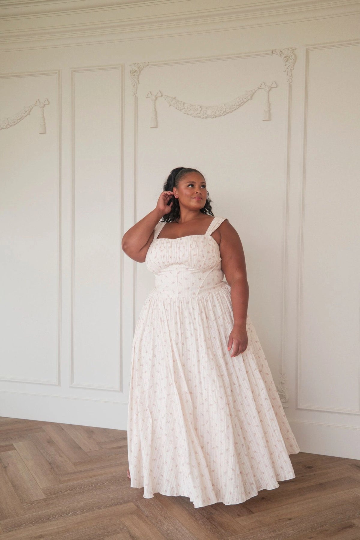 The image shows a woman in a white dress standing in front of a white wall, with a floor at the bottom. She is wearing a long, flowing dress with a high neckline and long sleeves, and her hair is