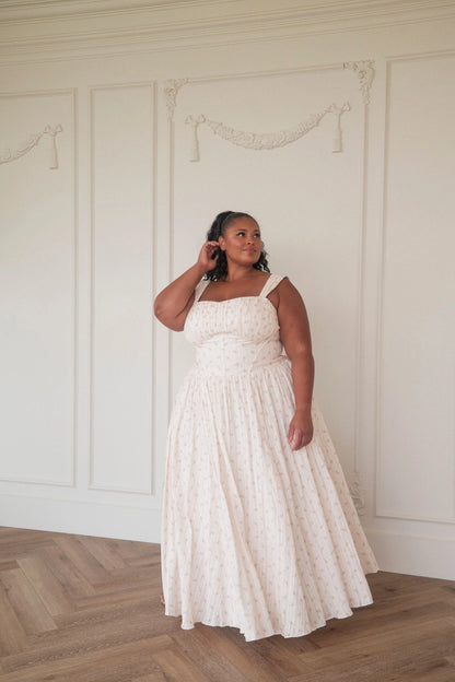 The image shows a woman in a white dress standing in front of a white wall, with a floor at the bottom. She is wearing a long, flowing dress with a high neckline and long sleeves, and her hair is