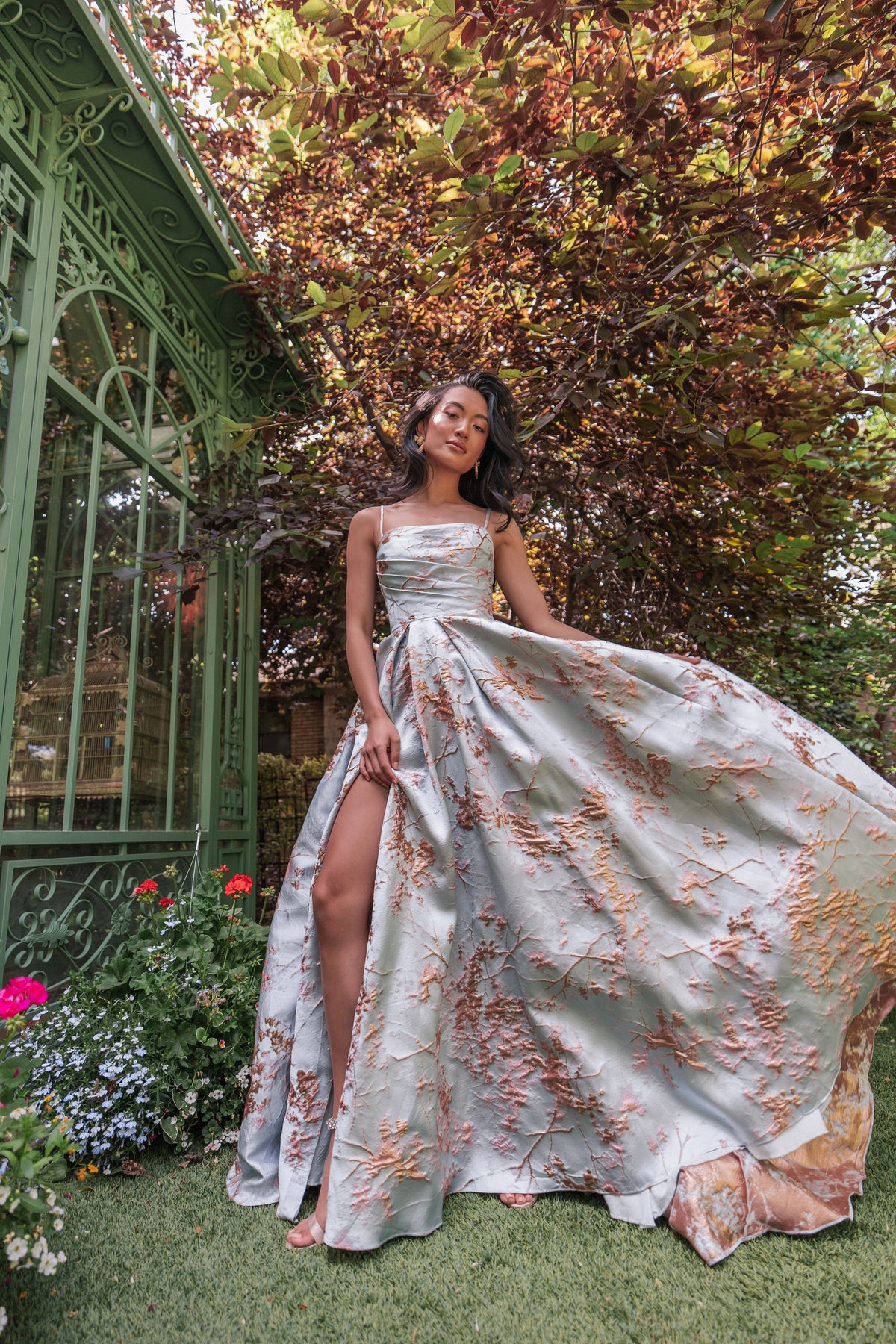 The image shows a woman in a long dress standing in front of a green archway surrounded by lush greenery and colorful flowers. The archway is adorned with intricate designs and the sky is visible in