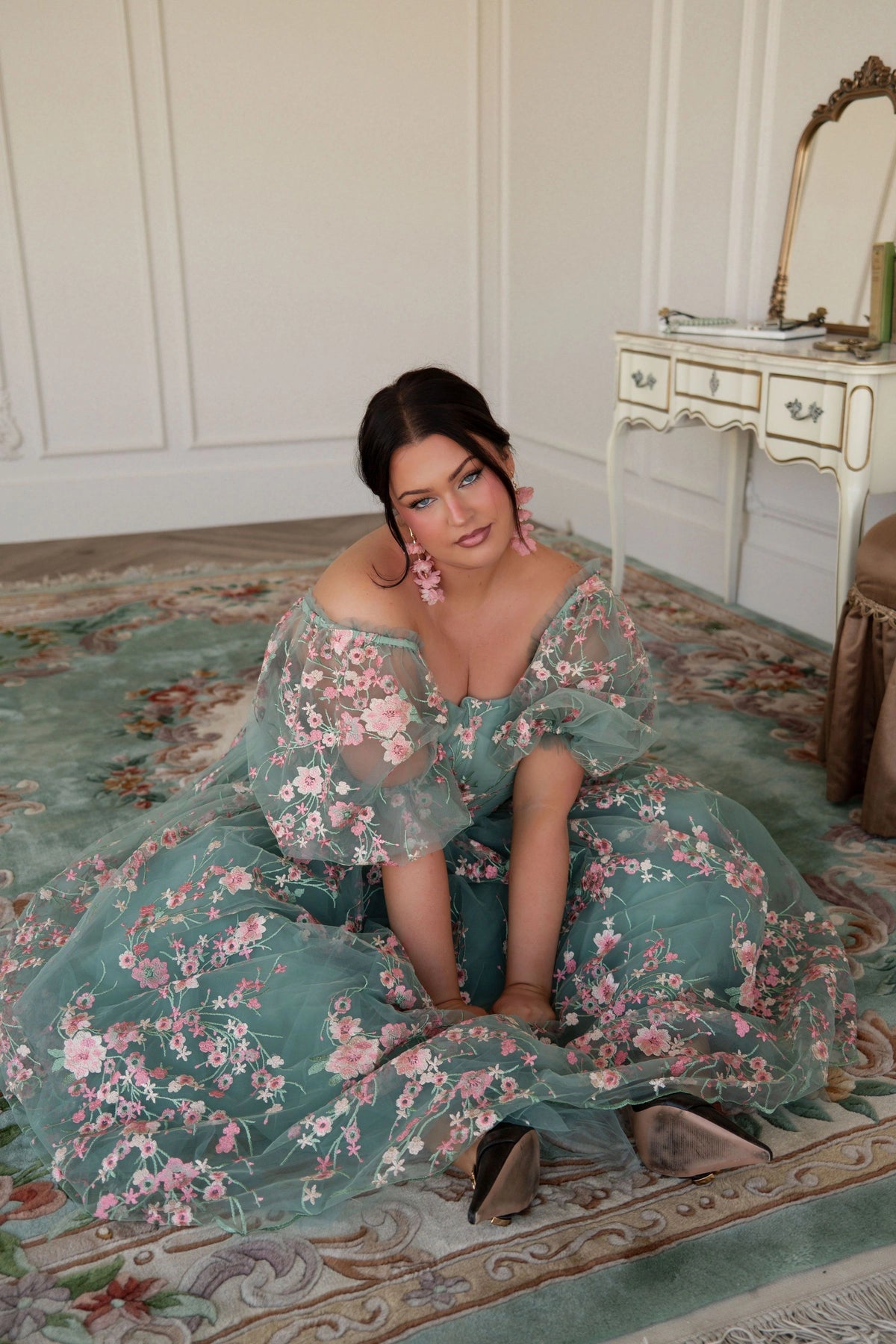 The image shows a woman in a green dress sitting on the floor in front of a mirror, with a table to her right and a wall in the background.