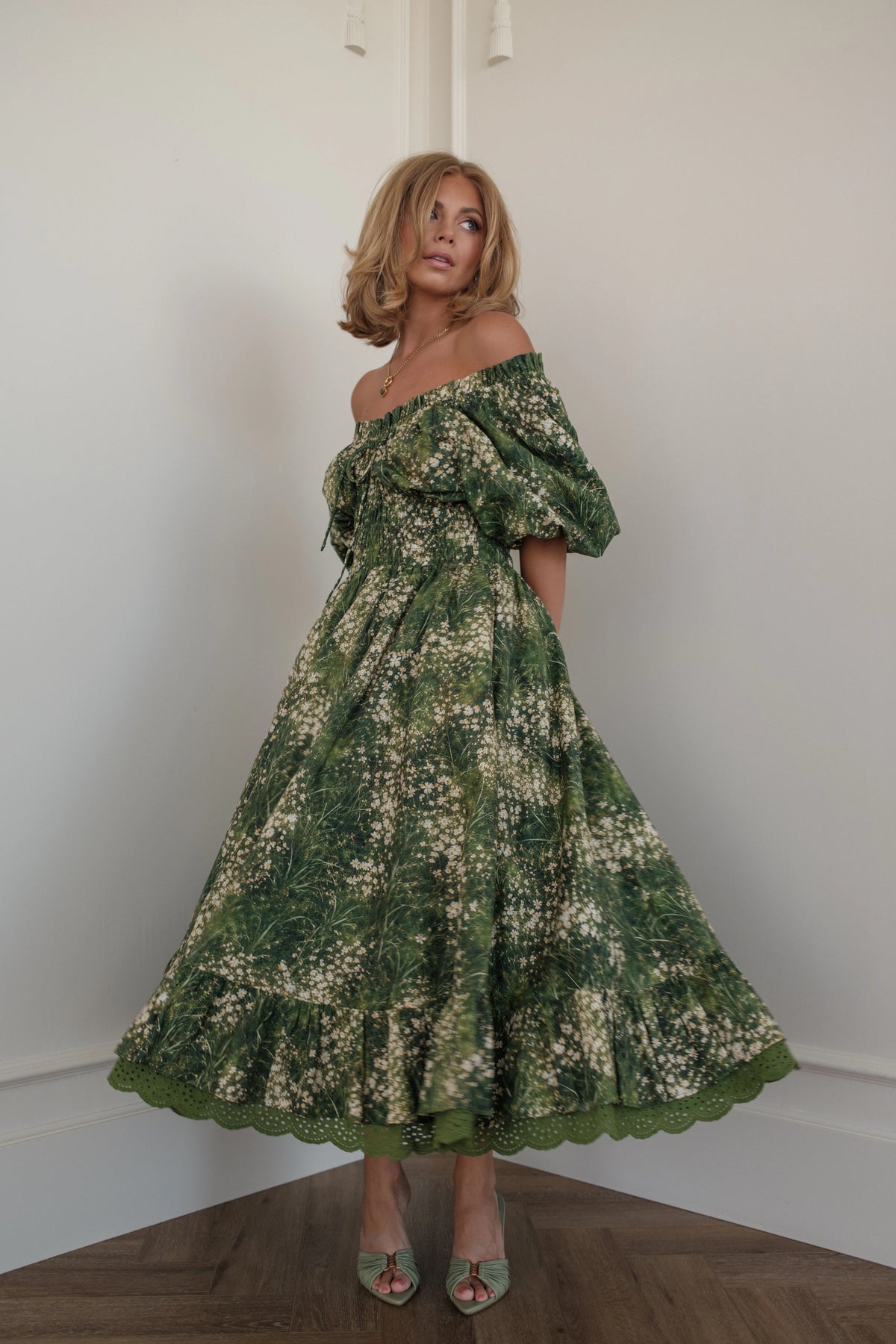 Woman in green floral off-the-shoulder dress and heels poses in corner of white room.