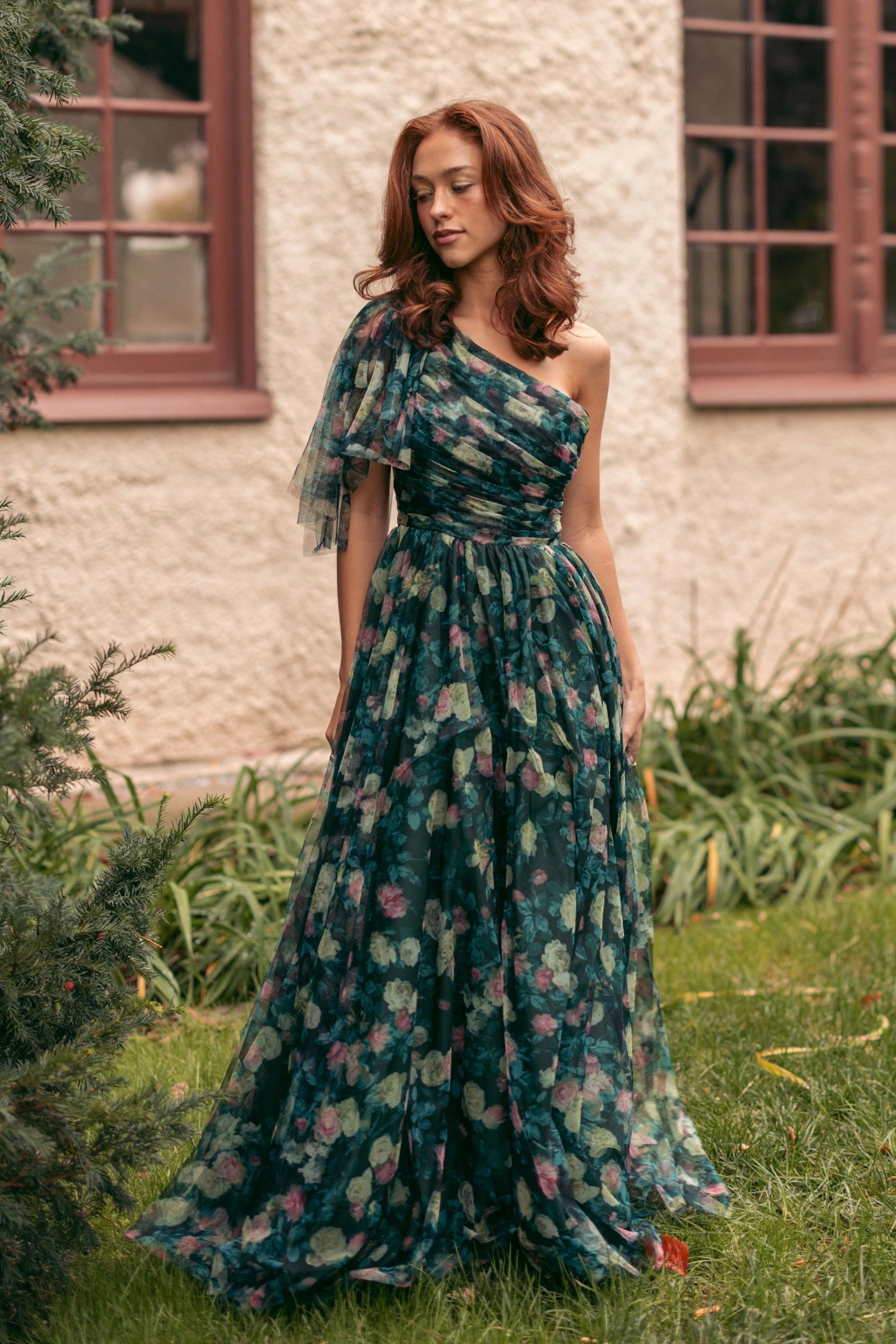 A woman in a green dress standing in front of a building with glass windows, surrounded by lush green grass and plants.
