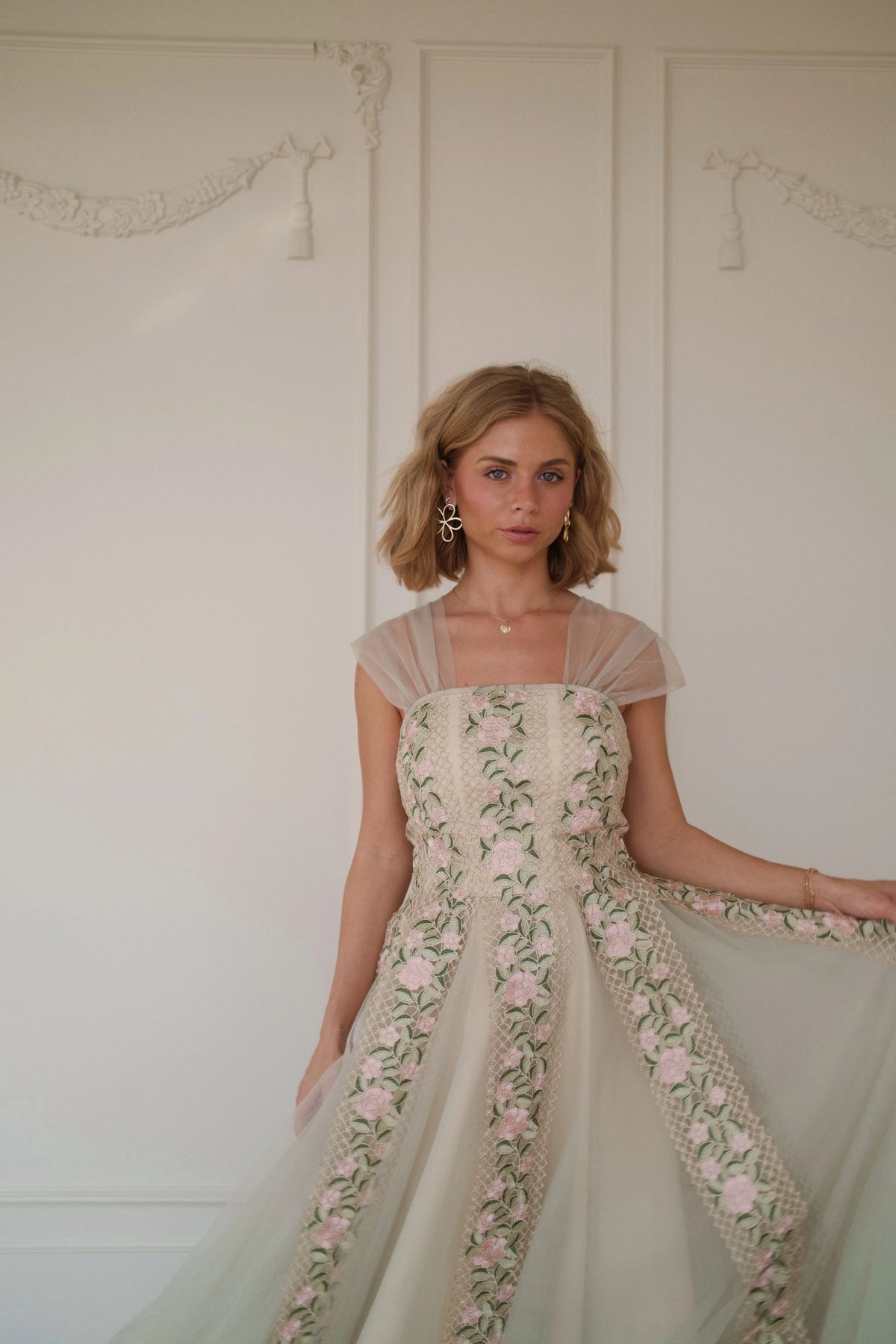 Woman in a white floral dress with short sleeves and a full skirt poses against a white wall.