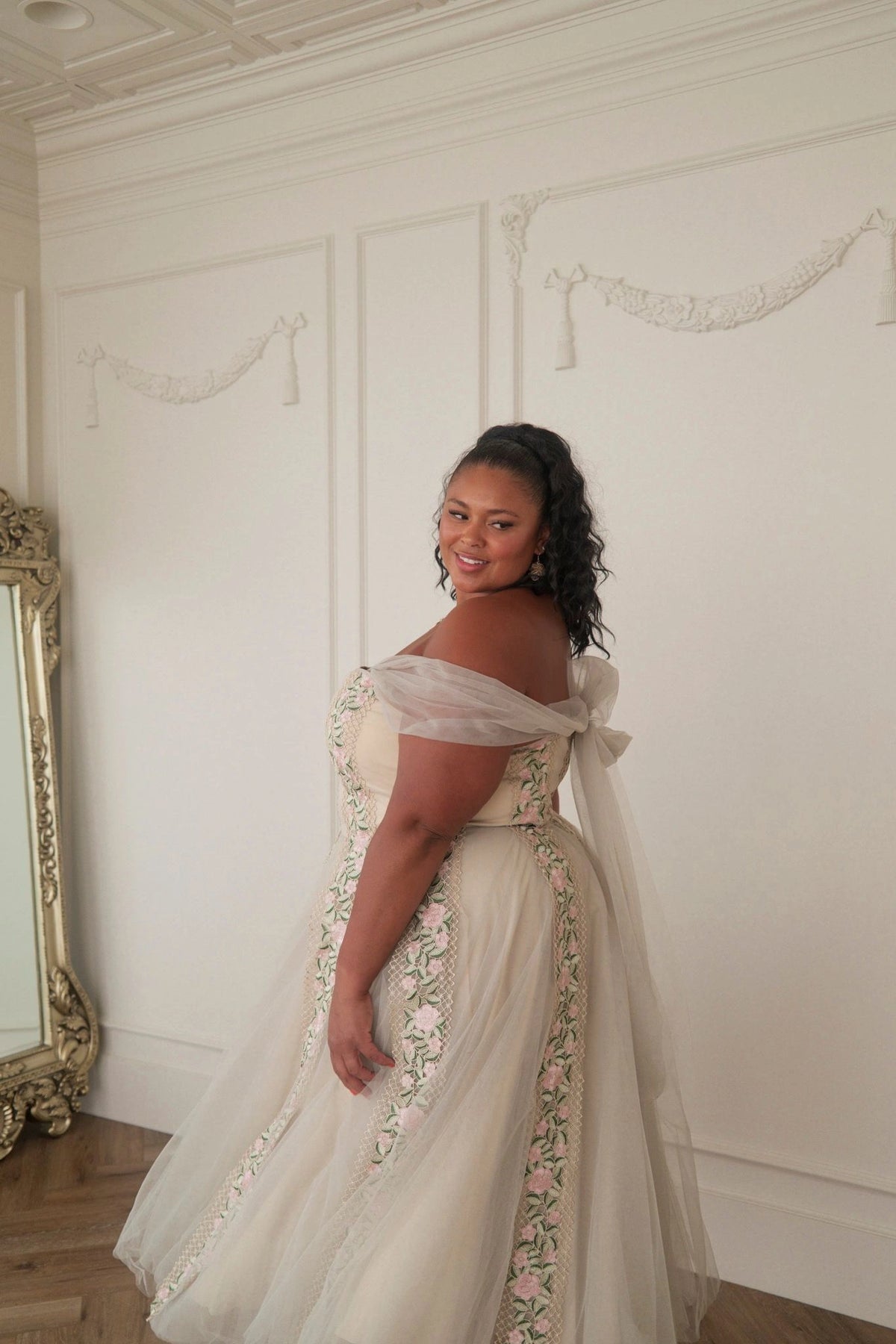 The image shows a woman in a white wedding dress standing in front of a mirror, with a wall in the background. She is wearing a white dress with intricate details, and her hair is styled in an