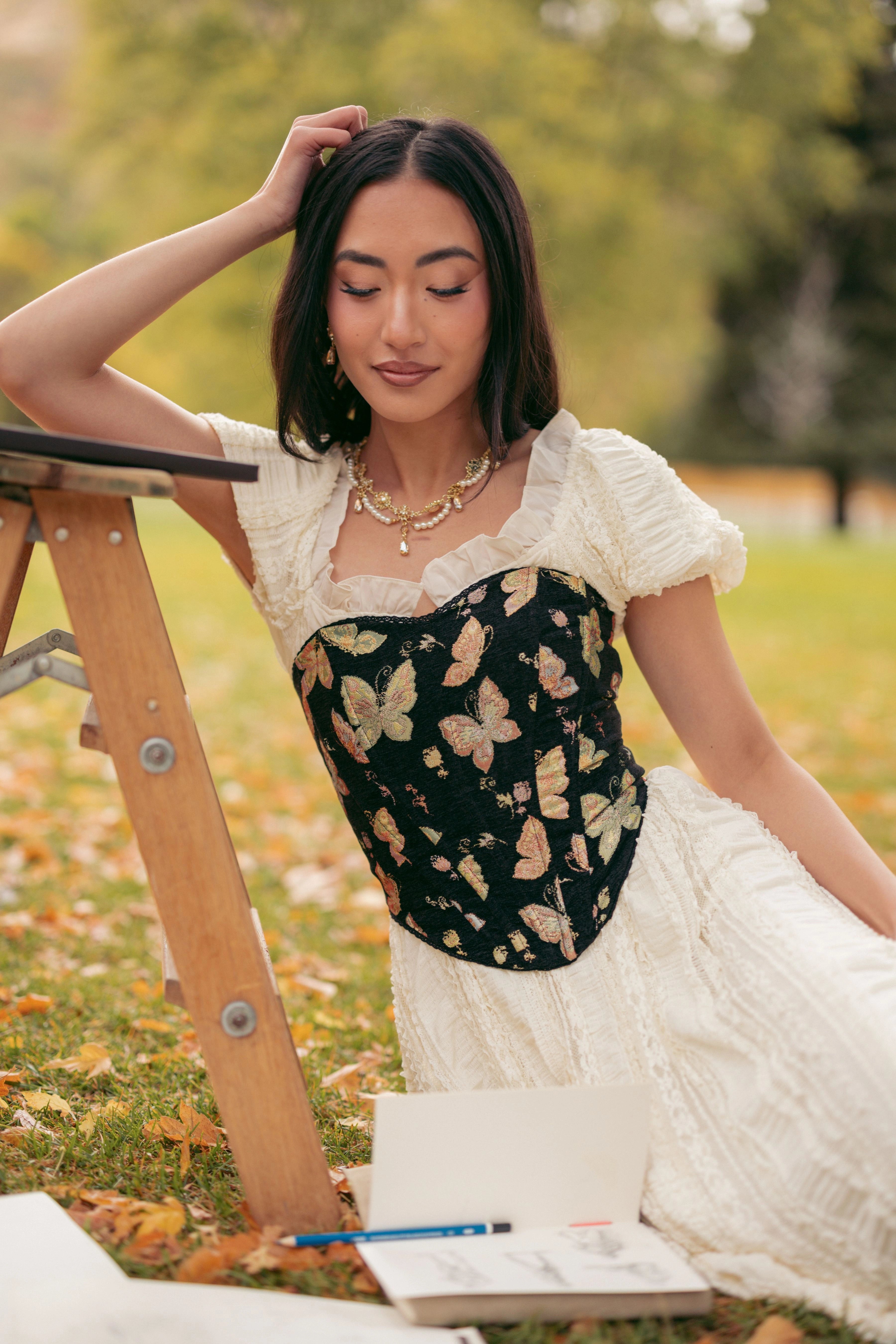 A woman in a white dress sitting on a wooden chair surrounded by trees and grass. She has a human face and is wearing a necklace.