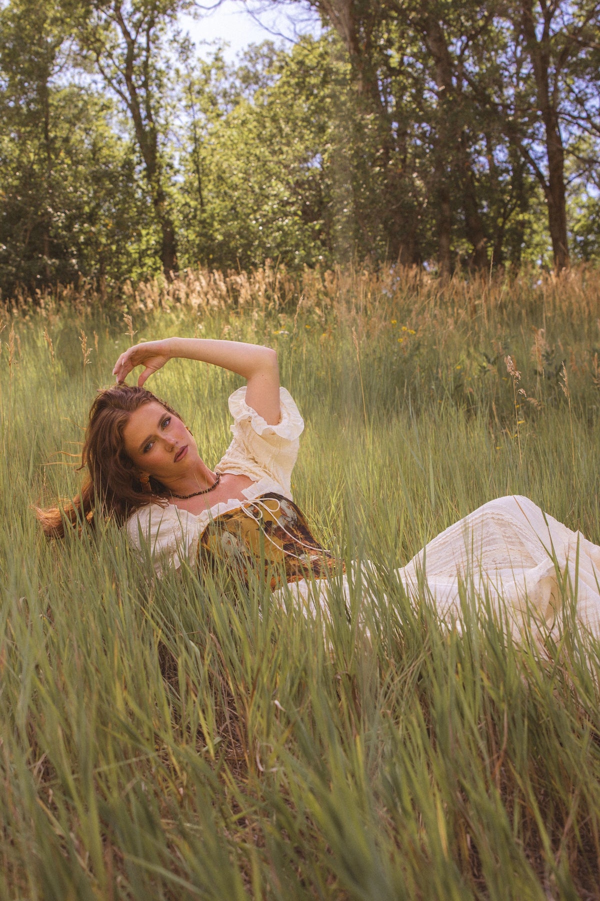 The image shows a woman in a white dress laying in a field of tall grass, surrounded by trees and a clear blue sky.