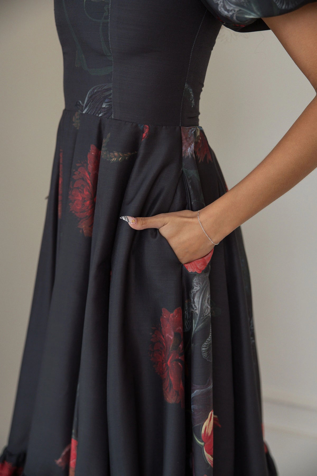 The image shows a woman wearing a black dress with a red and black floral print, standing in front of a wall. The dress is a long, flowing silhouette with a high neckline and long sleeves, and the