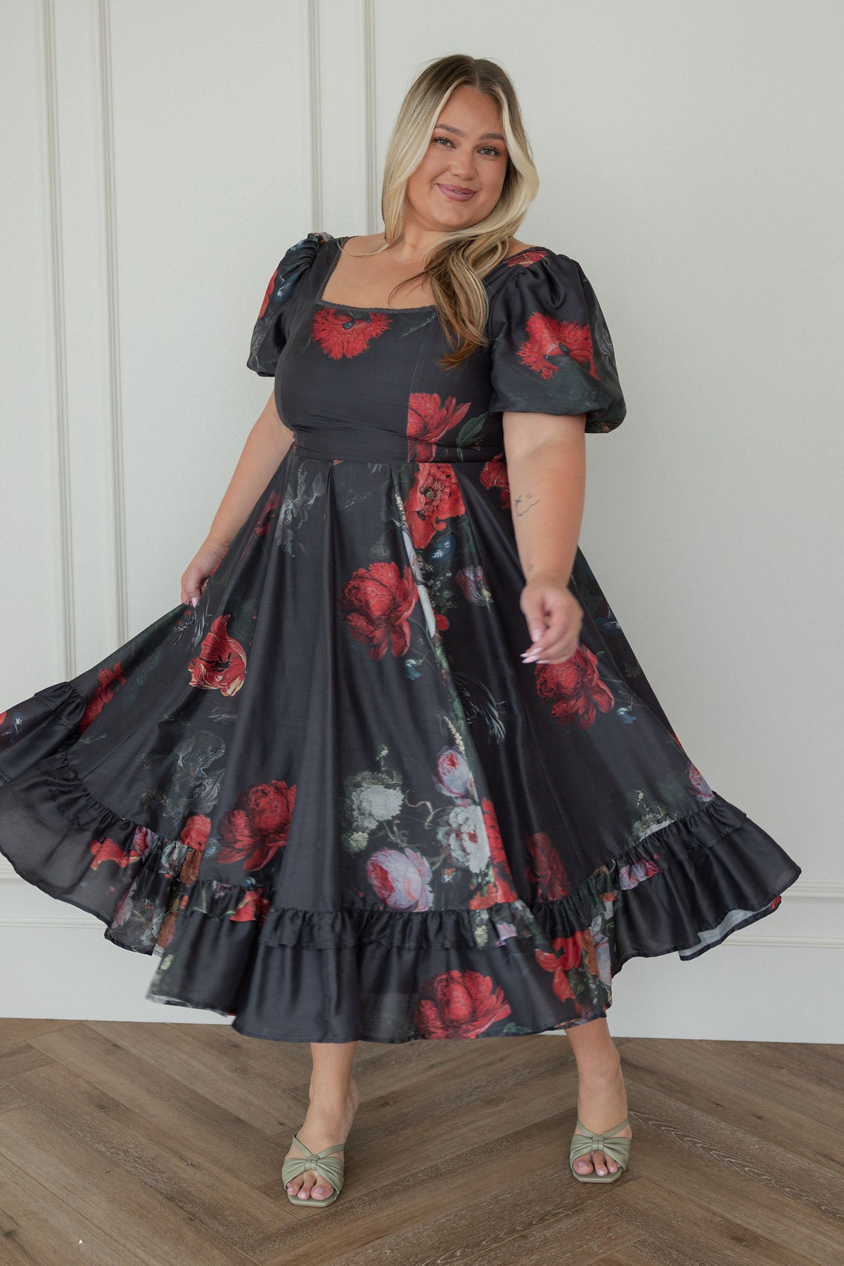 The image shows a woman standing in front of a white wall wearing a black and red floral dress. The dress has a unique pattern of red roses, and the woman is standing on a wooden floor.