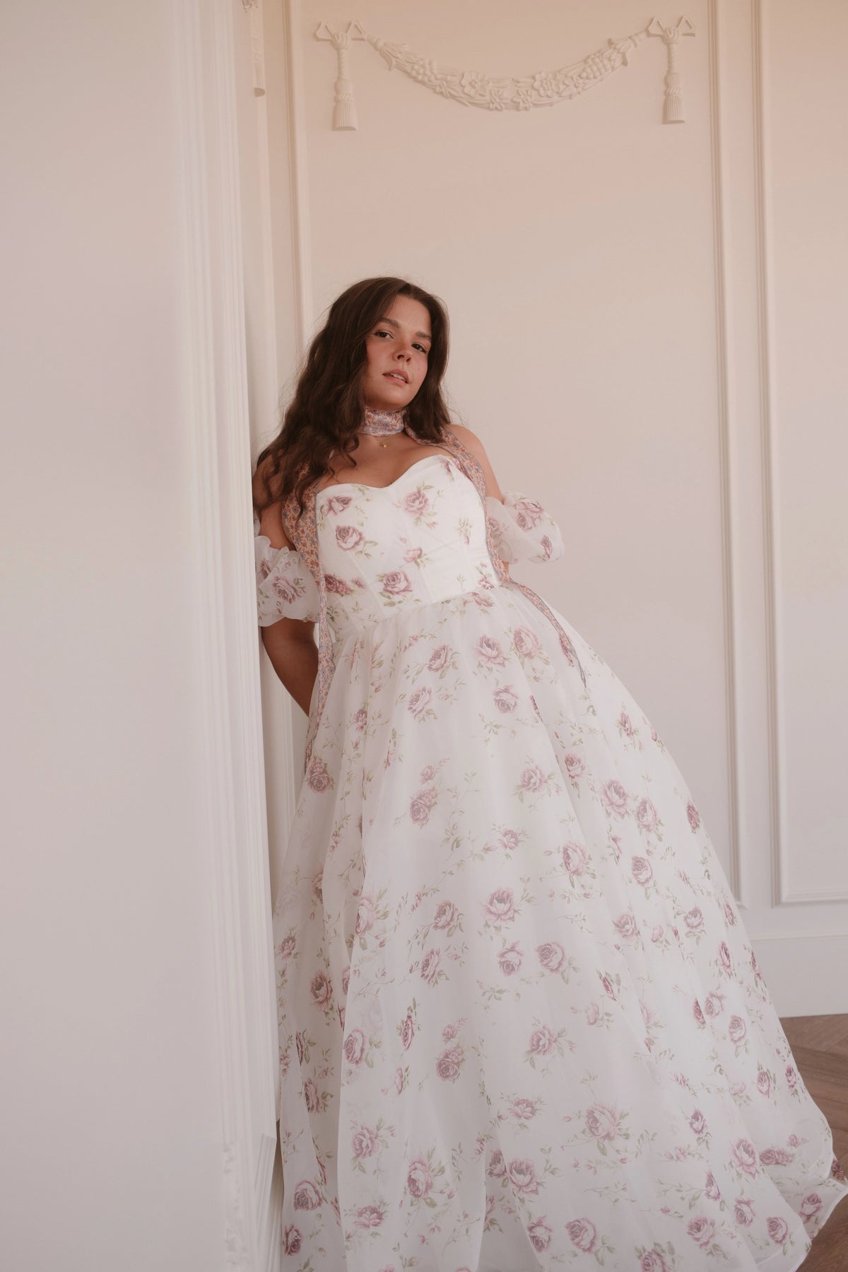 Woman in white floral dress with pink roses and lace details leaning against a doorway, looking to the right.