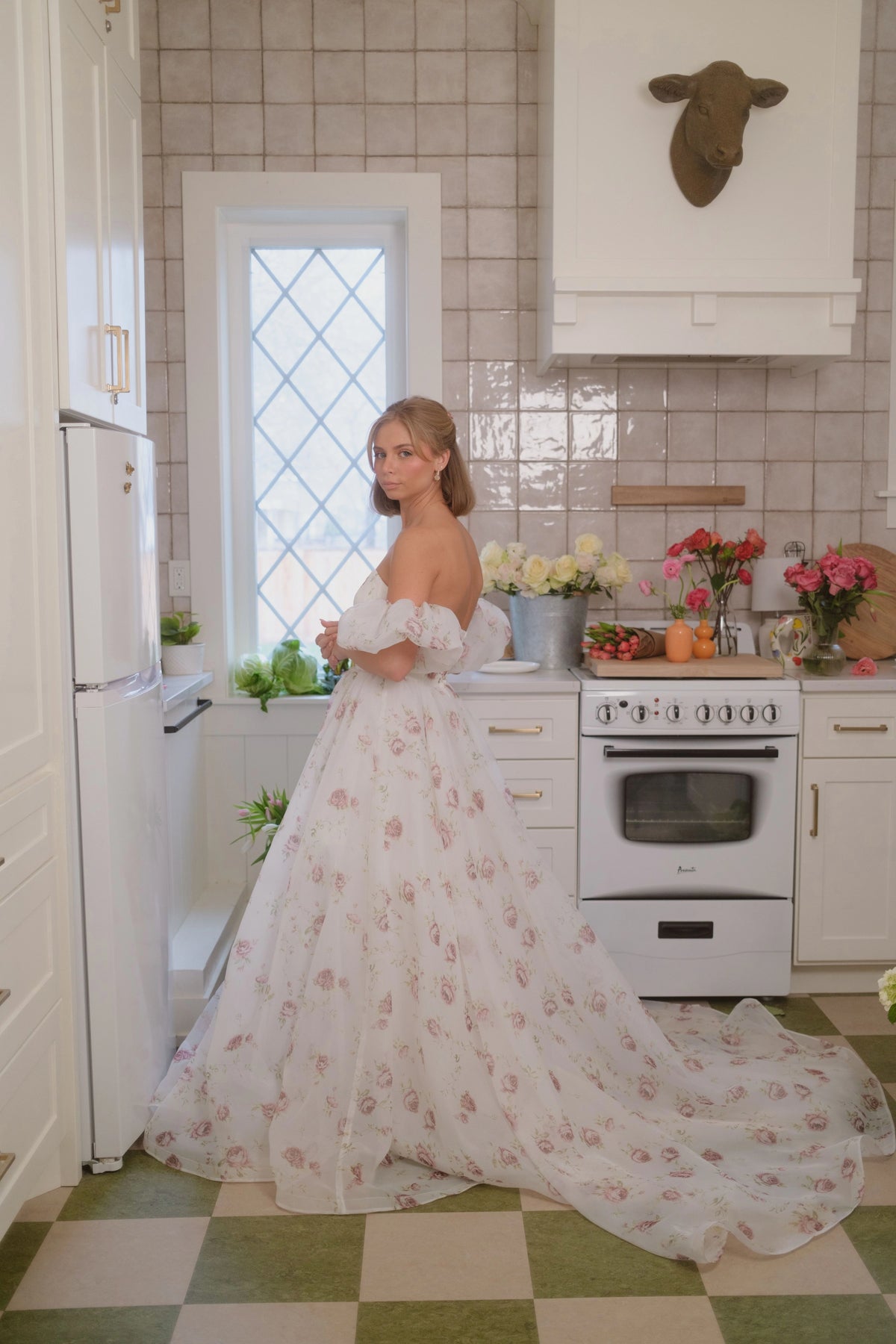 Woman in white floral wedding dress with off-shoulder sleeves stands in a kitchen with white appliances and green checkered floor.
