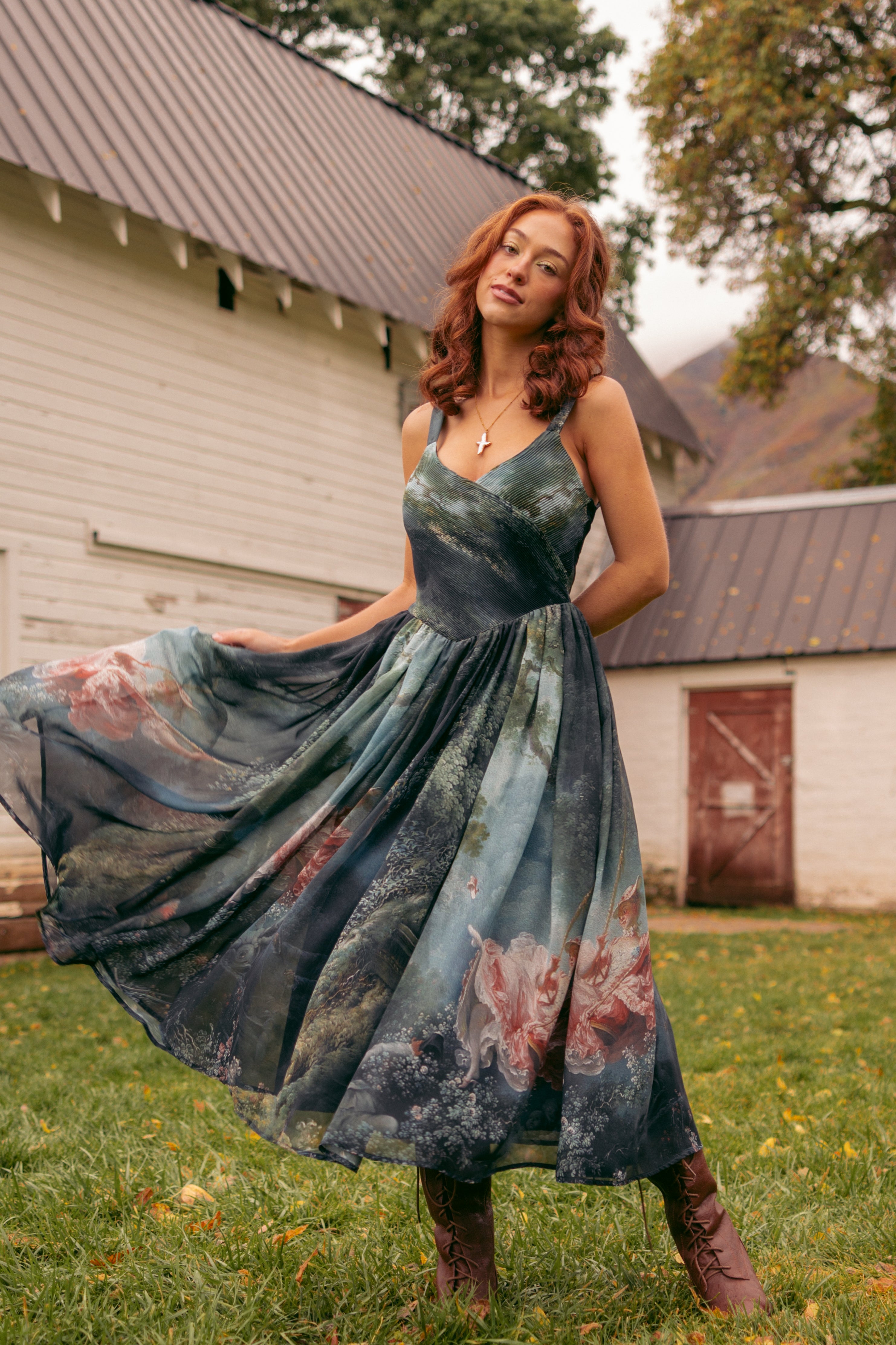 A woman in a long dress standing in the grass in front of a house, surrounded by trees and a clear blue sky.