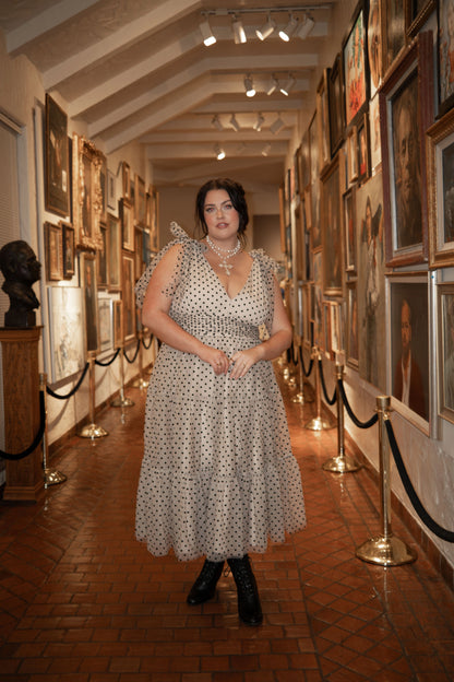 A woman in a white dress walking down a hallway lined with photo frames on the walls and illuminated by lights at the top.