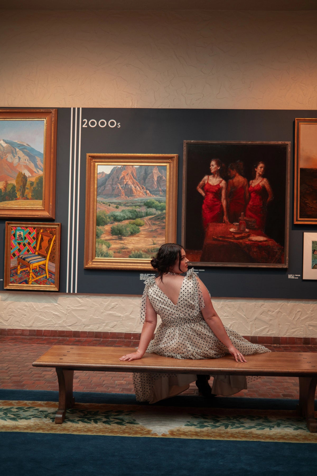A woman sitting on a bench in front of a wall adorned with paintings. She is wearing a white dress and has her hair pulled back. The wall behind her is filled with a variety of paintings, each one