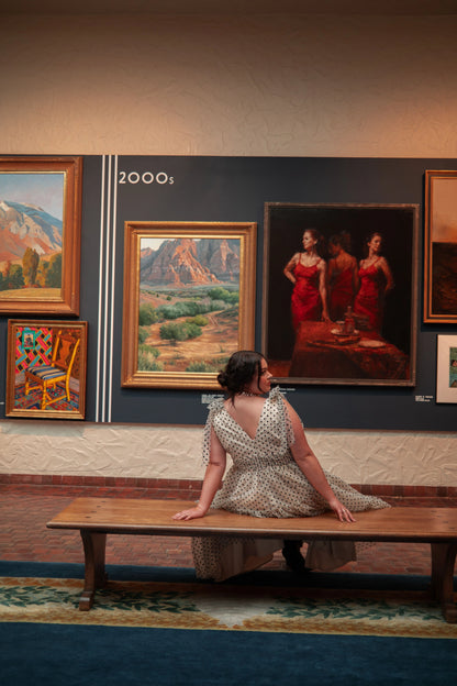 A woman sitting on a bench in front of a wall adorned with paintings. She is wearing a white dress and has her hair pulled back. The wall behind her is filled with a variety of paintings, each one