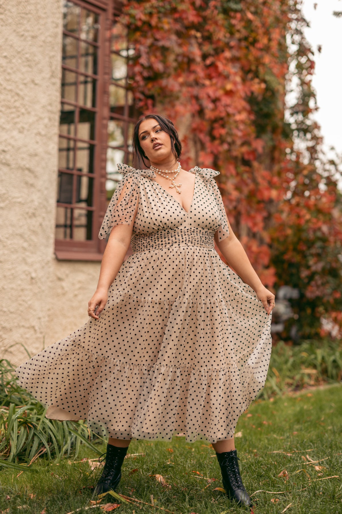 A woman in a polka dot dress walking through a lush green field, with a wall and window in the background. The sky is visible above, and the woman is surrounded by plants and trees.