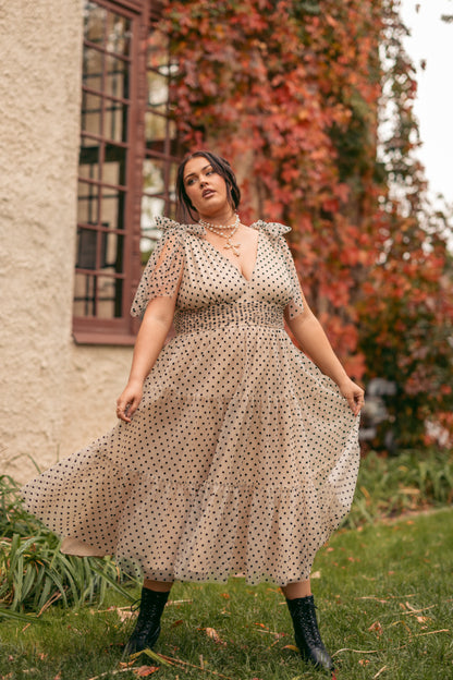A woman in a polka dot dress walking through a lush green field, with a wall and window in the background. The sky is visible above, and the woman is surrounded by plants and trees.