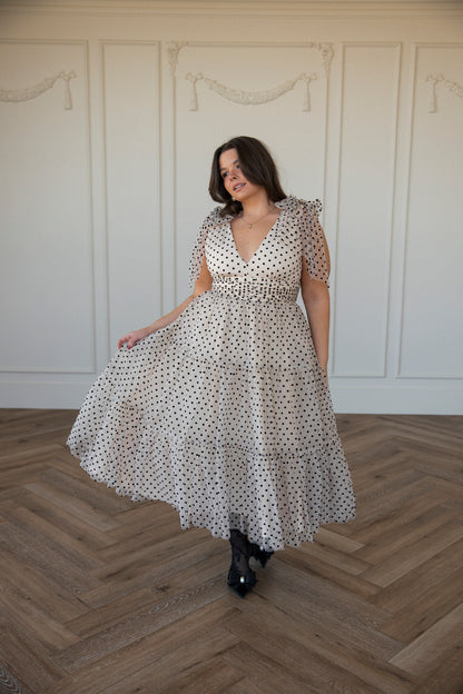The image shows a woman in a white polka dot dress standing on a wooden floor, with a wall in the background. She is wearing a dress with a unique pattern of black and white polkas, giving her a