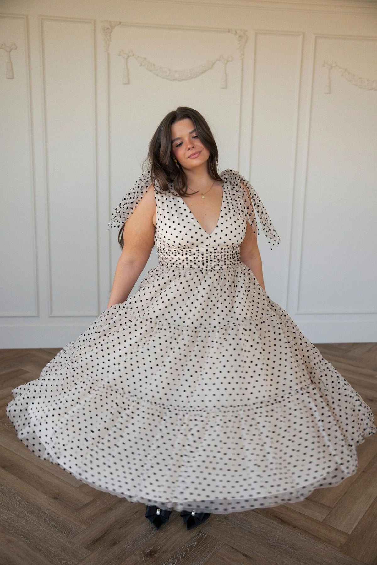 The image shows a woman in a white dress standing on a wooden floor, with a wall in the background. She is wearing a long, flowing dress with a high neckline and long sleeves, and her hair is styled