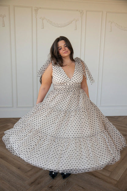 The image shows a woman in a white dress standing on a wooden floor, with a wall in the background. She is wearing a long, flowing dress with a high neckline and long sleeves, and her hair is styled