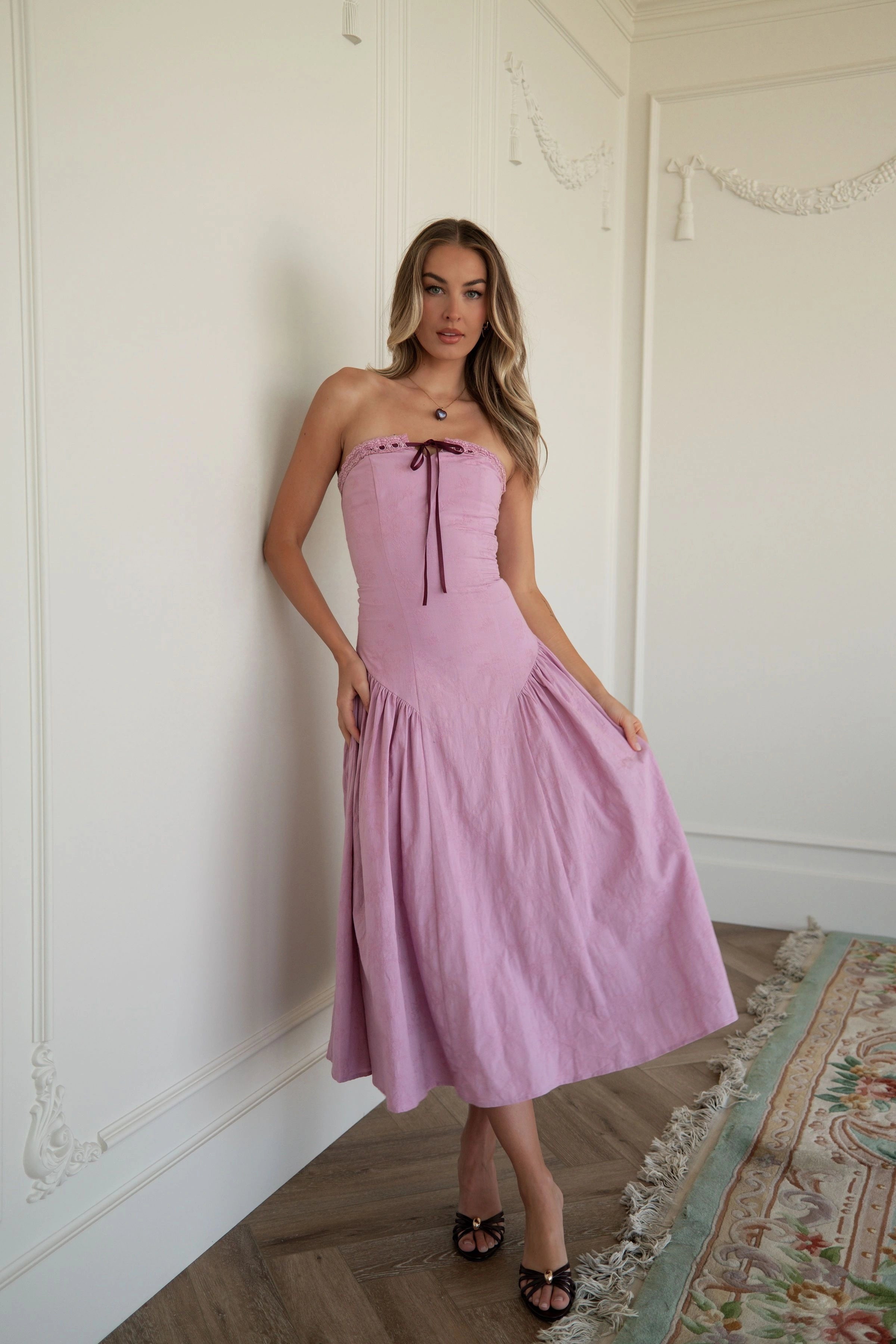 The image shows a woman standing in front of a white wall wearing a pink dress. She is standing on a carpeted floor, and the dress is a light pink color.