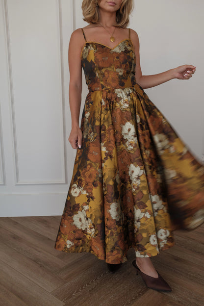 Woman in a brown, yellow and white floral maxi dress with spaghetti straps and brown heels poses against a white wall.