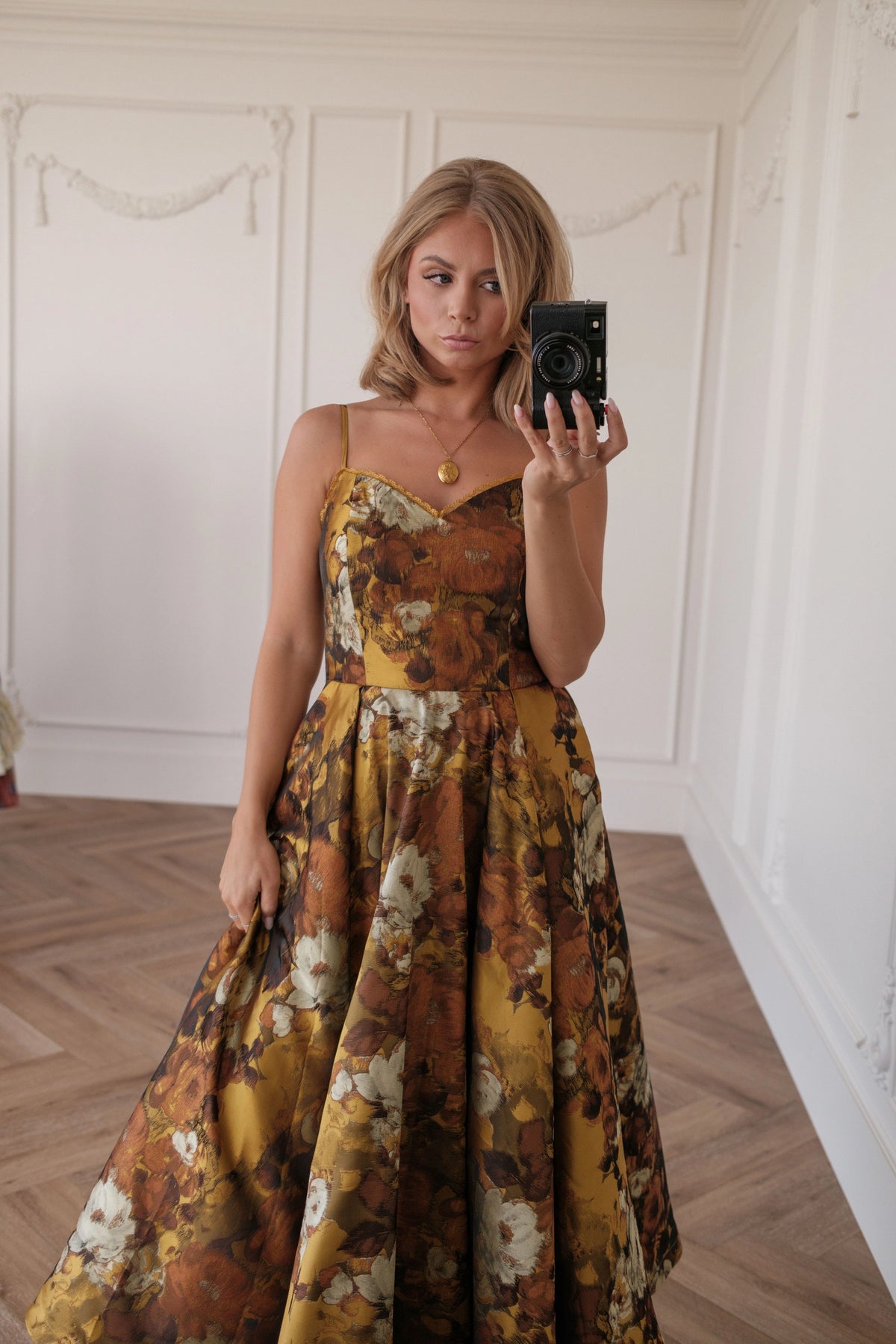 Woman in a yellow floral maxi dress takes selfie with black camera in a white room.