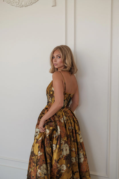 Woman in a brown floral dress with spaghetti straps poses against a white wall, looking over her shoulder.