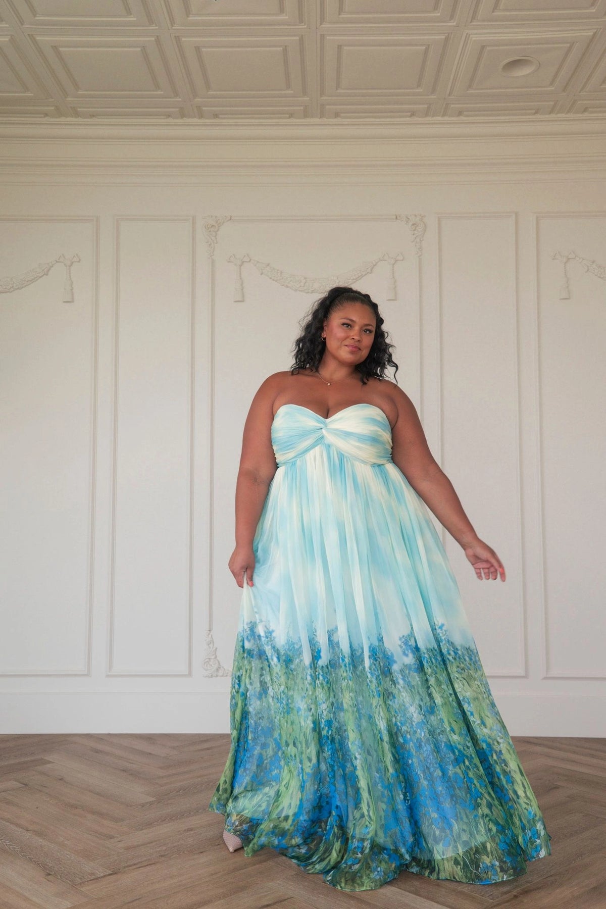 The image shows a woman standing in front of a white wall wearing a beautiful blue and green dress. The dress is made of a light fabric and has a fitted bodice with a full skirt that falls to the