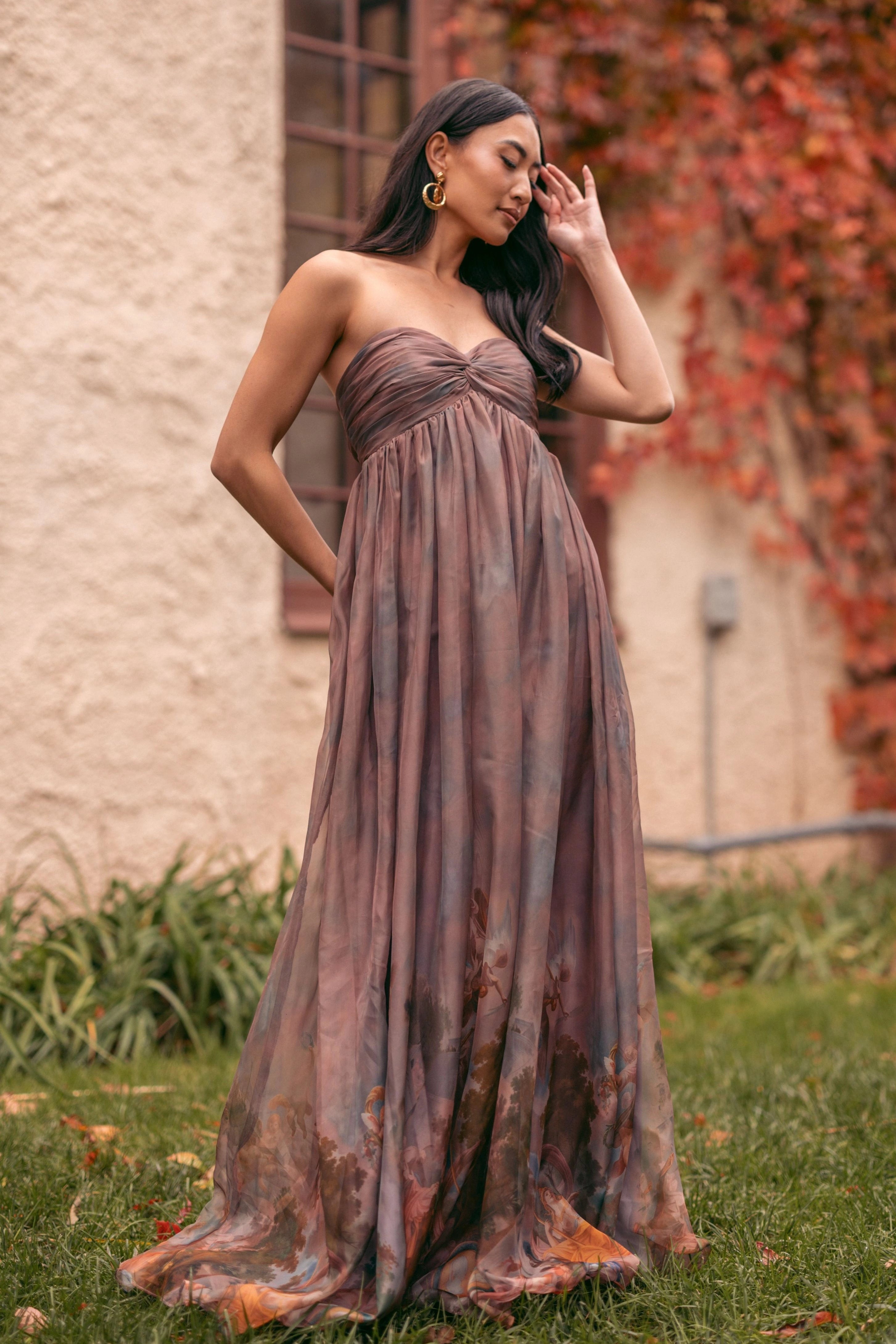 A woman in a long dress standing in front of a building with a window and a wall, surrounded by plants and grass.