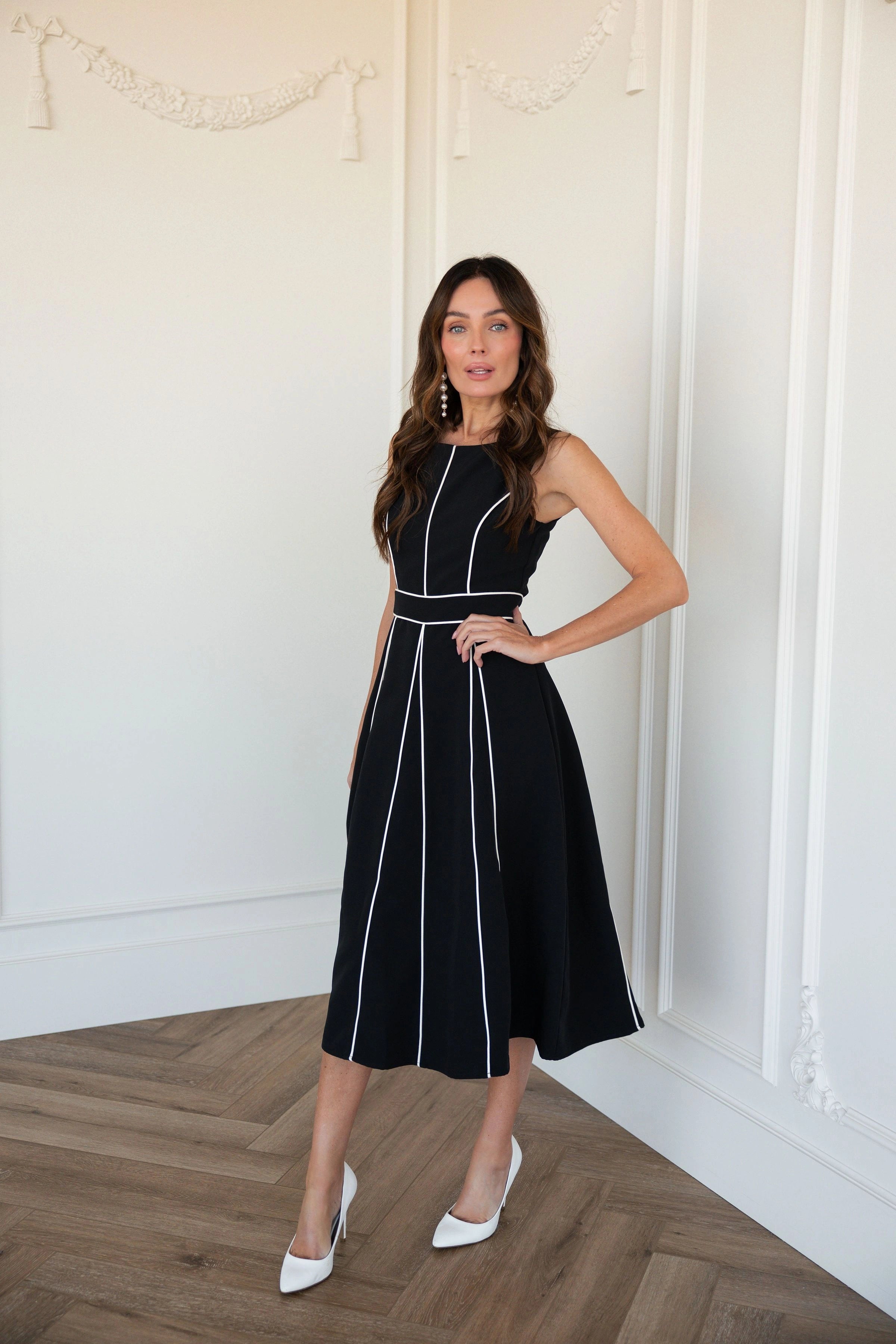 The image shows a woman standing in front of a white wall wearing a black and white dress. She is standing on the floor, with the wall behind her.