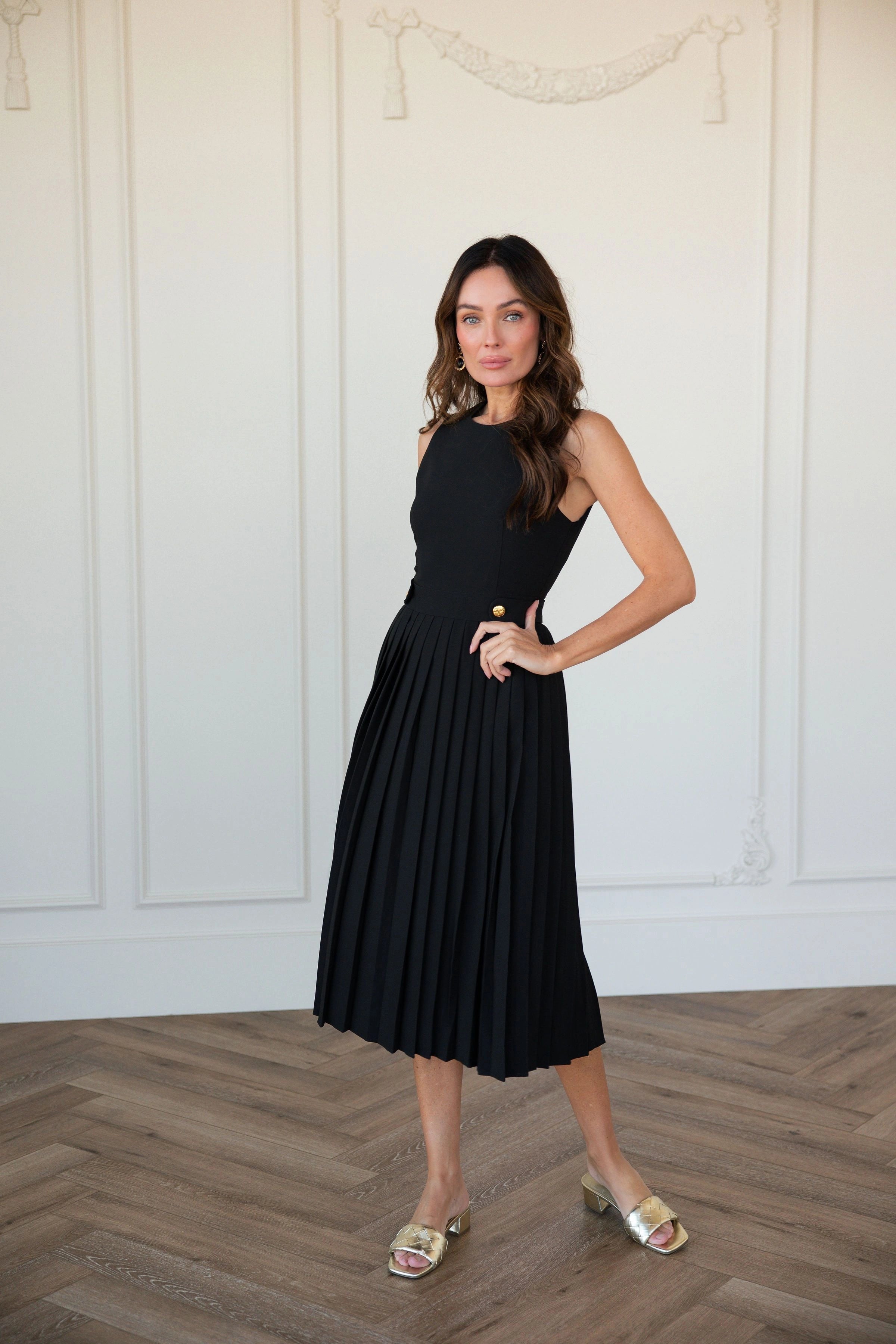The image shows a woman in a black dress standing in front of a white wall, posing for a picture. She is wearing a black frock and her hair is styled in a sleek bob. Her makeup is subtle and natural