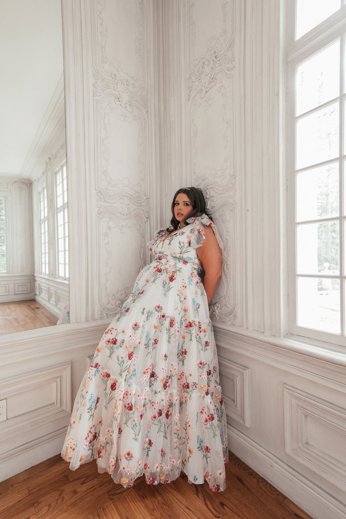 Woman in white floral maxi dress leaning against ornate white wall beside window.