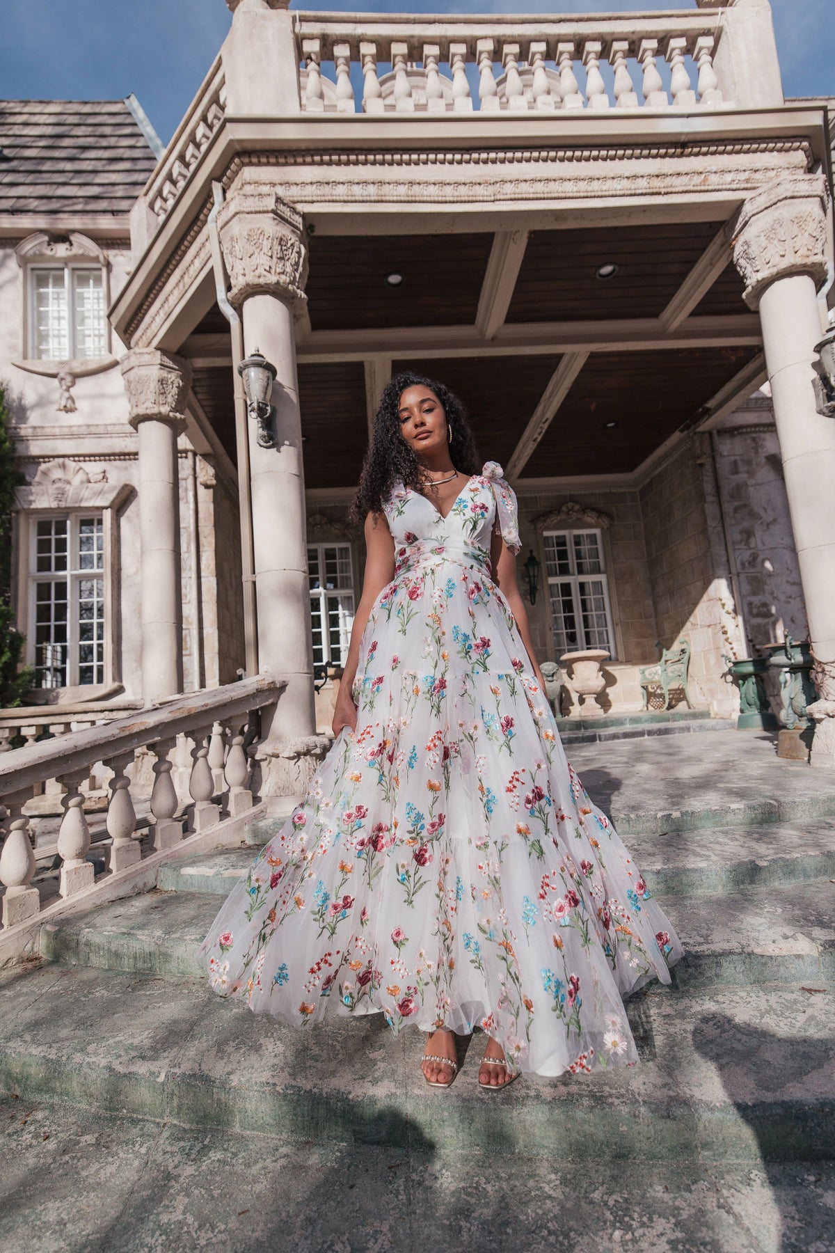 A woman in a white floral gown with short puffed sleeves poses on stone steps in front of a large ornate house.