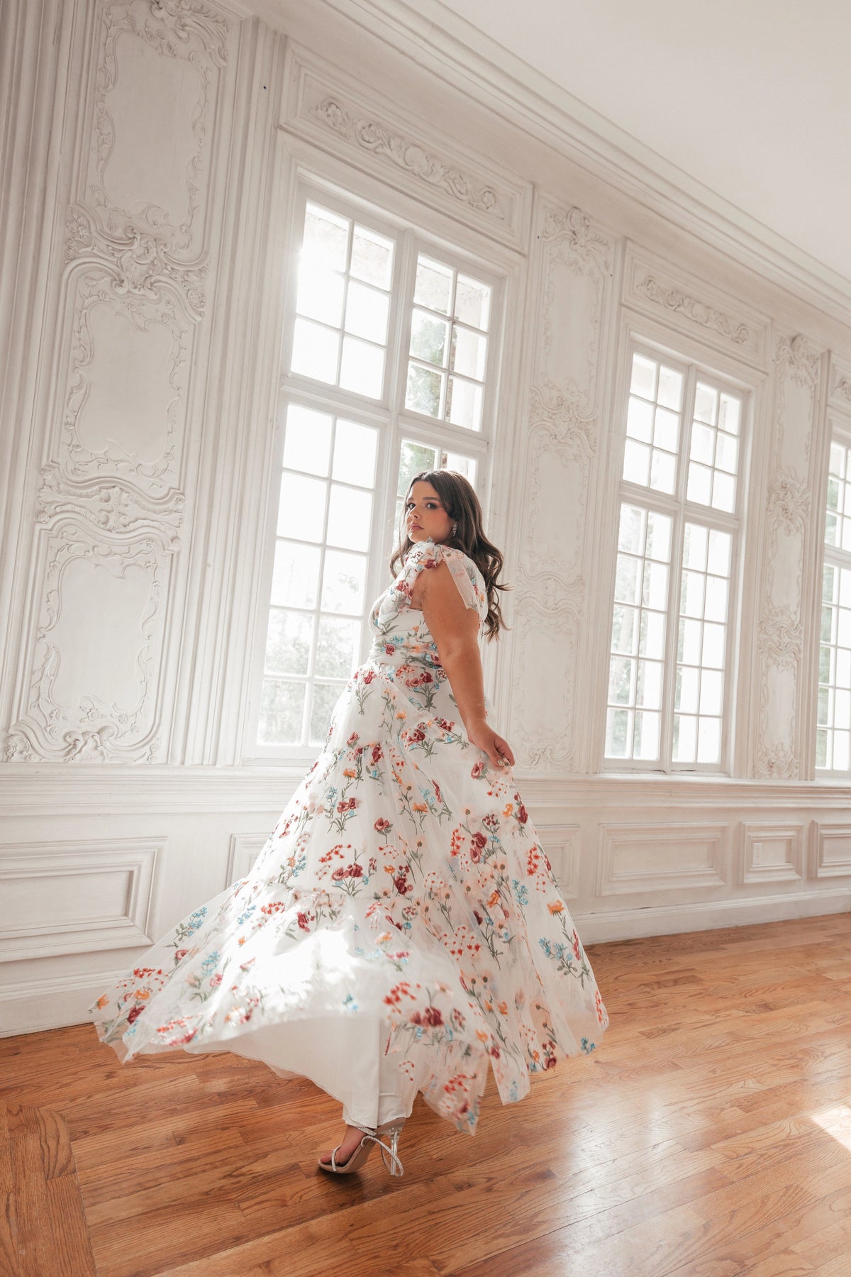 Woman in white floral dress with lace cap sleeves twirls in ornate room with large windows, wooden floor, and white walls.