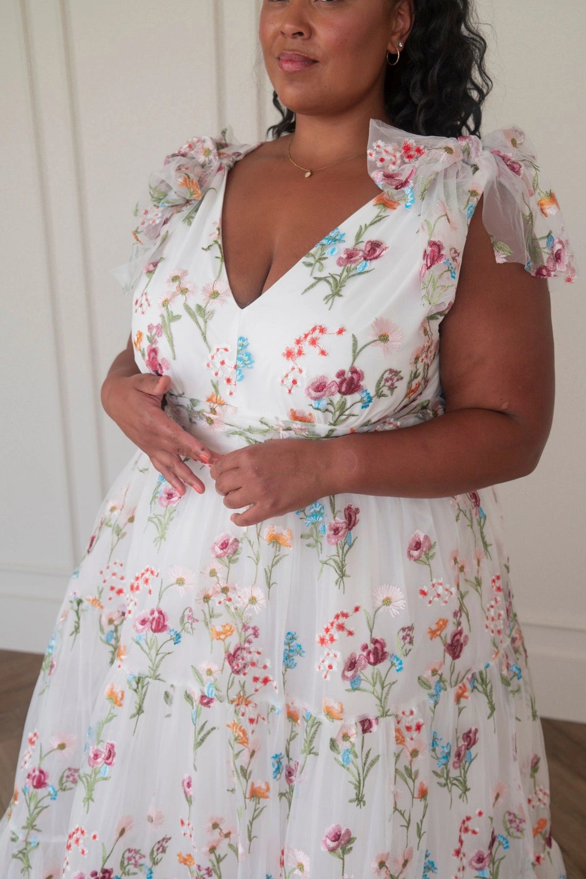 The image shows a woman in a white dress standing in front of a mirror, with a wall in the background. She is wearing a white frock with a floral pattern, and her hair is styled in a sleek bob. Her