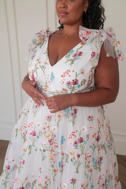 The image shows a woman in a white dress standing in front of a mirror, with a wall in the background. She is wearing a white frock with a floral pattern, and her hair is styled in a sleek bob. Her
