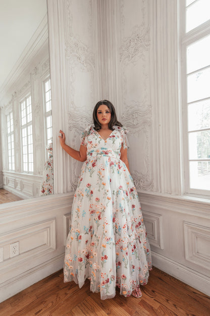 Woman in white floral maxi dress with V-neck and short sleeves poses beside a mirror in an ornate white room.