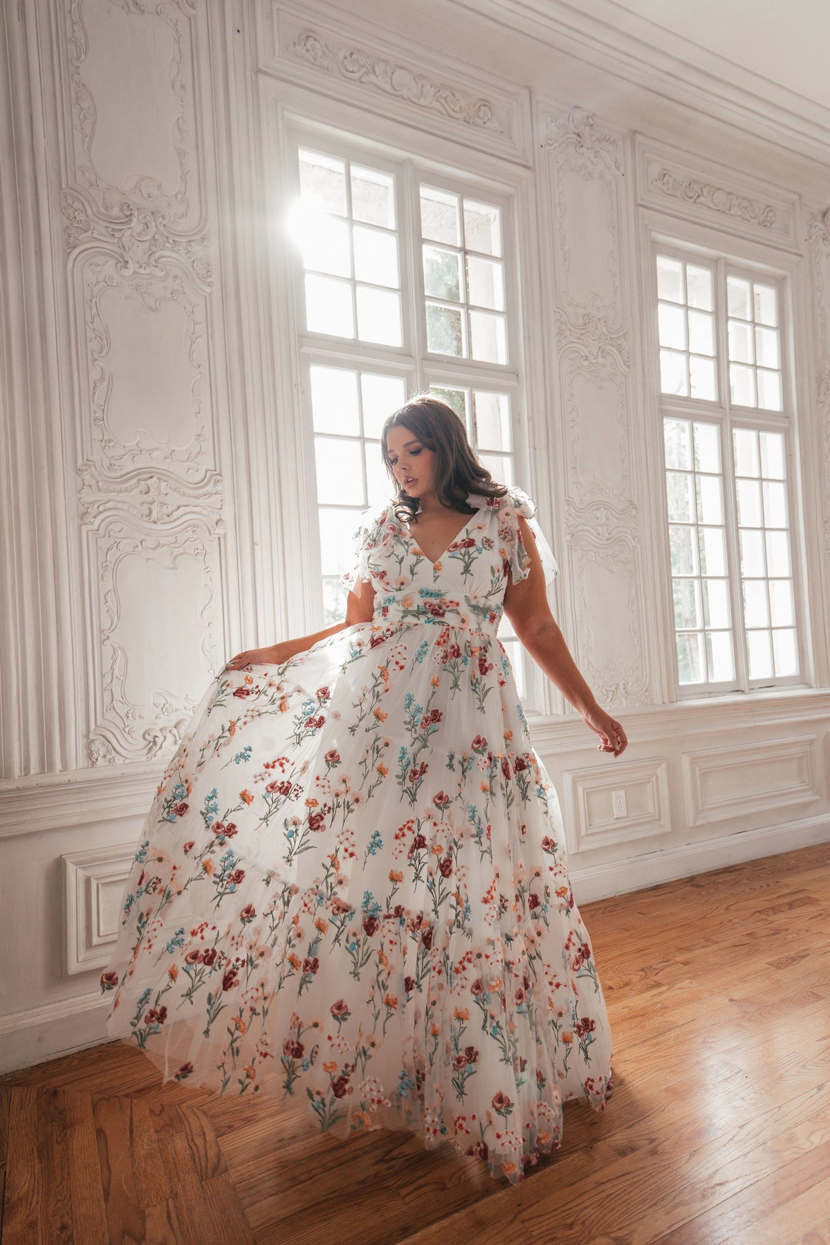 Woman in white floral maxi dress with V-neck and short sleeves poses in ornate room with large windows.