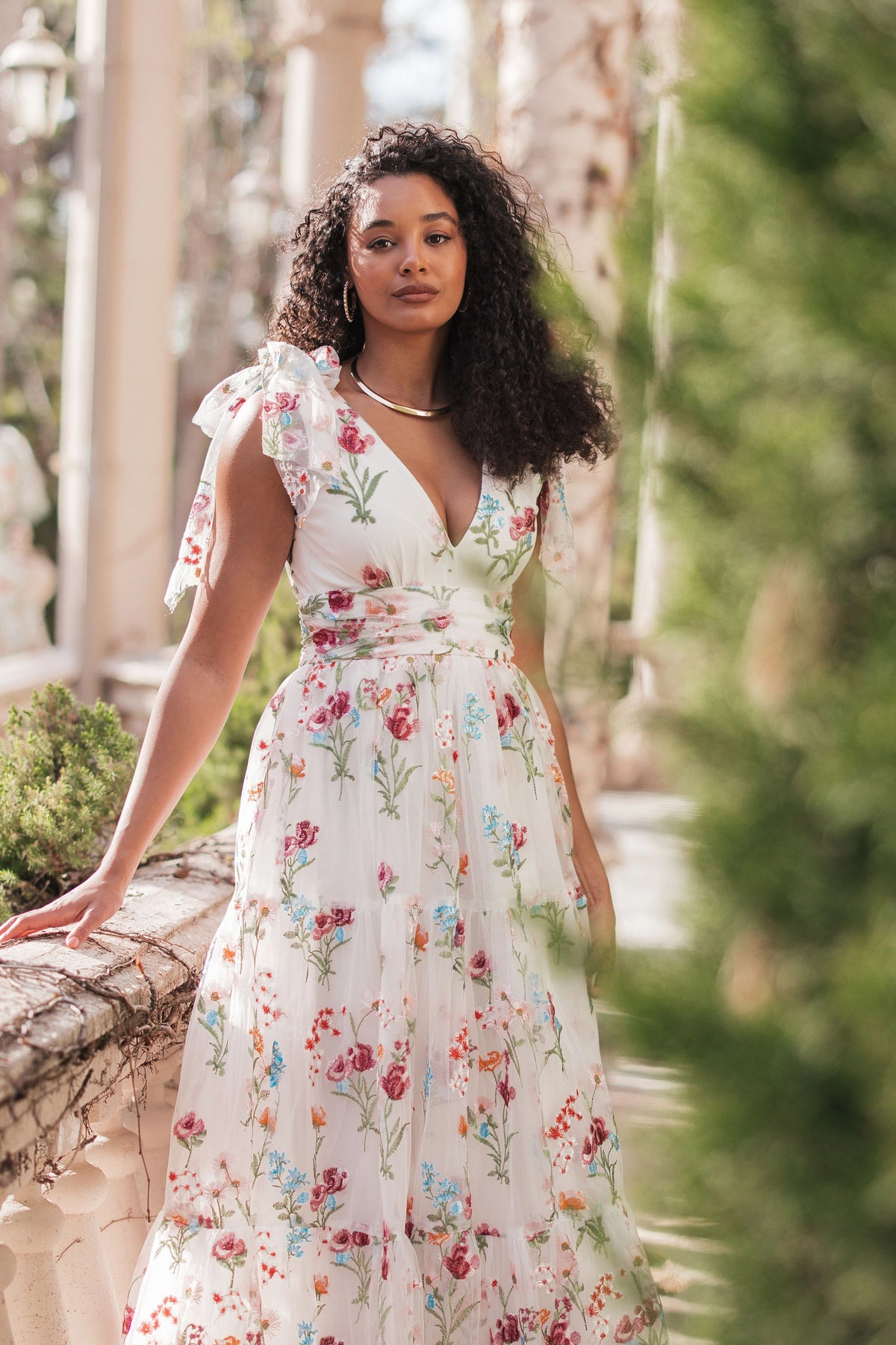 Woman in white floral dress with V-neck and bow sleeves poses beside a stone railing in a garden setting.