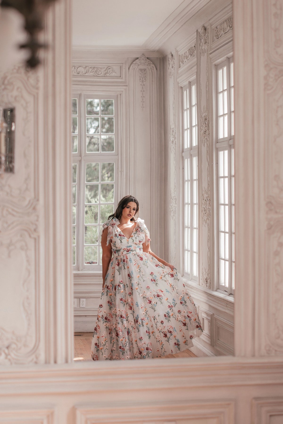 Woman in white floral dress with ruffled sleeves stands in ornate room with large windows.