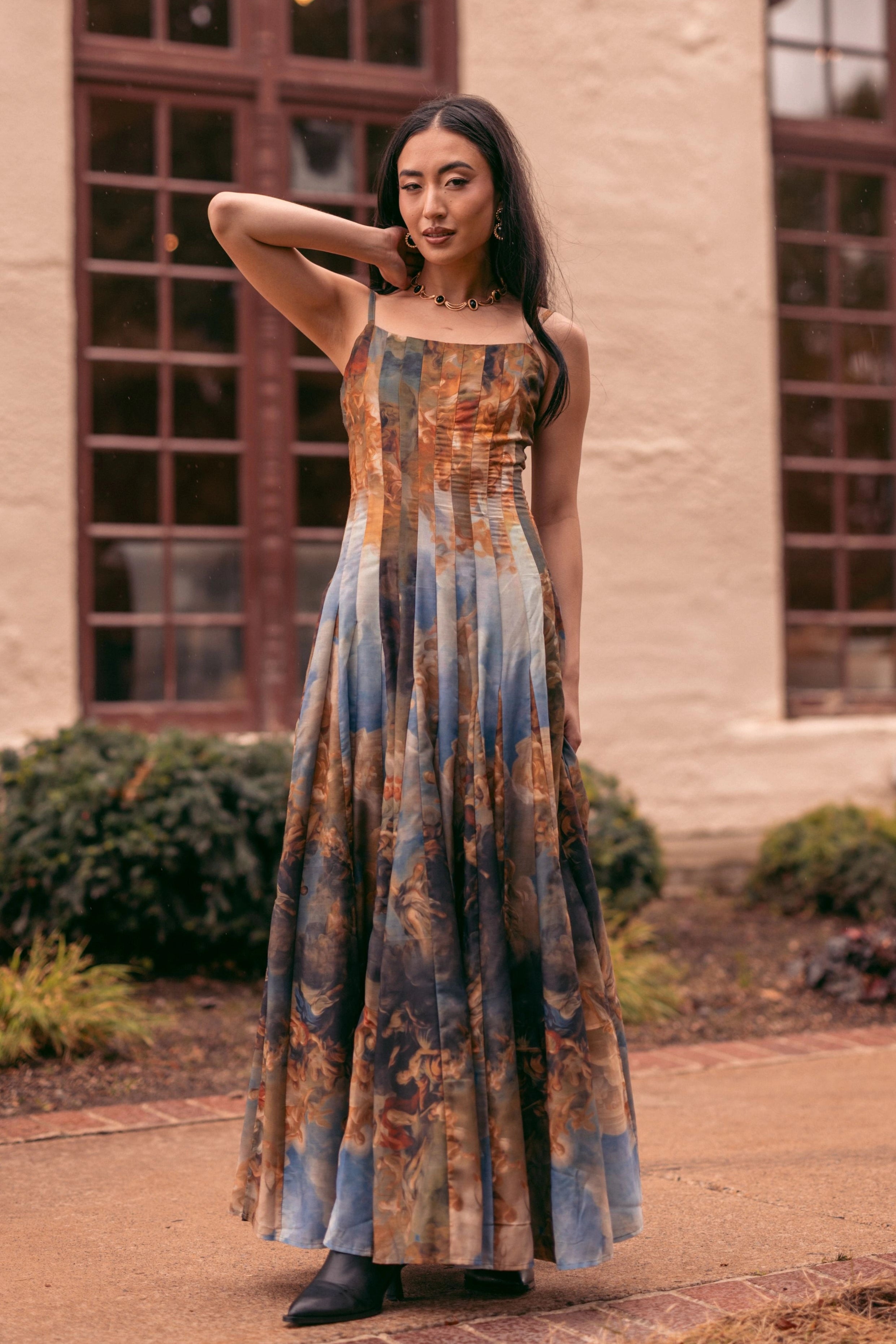 A woman in a long dress standing in front of a building with windows, surrounded by plants and grass.