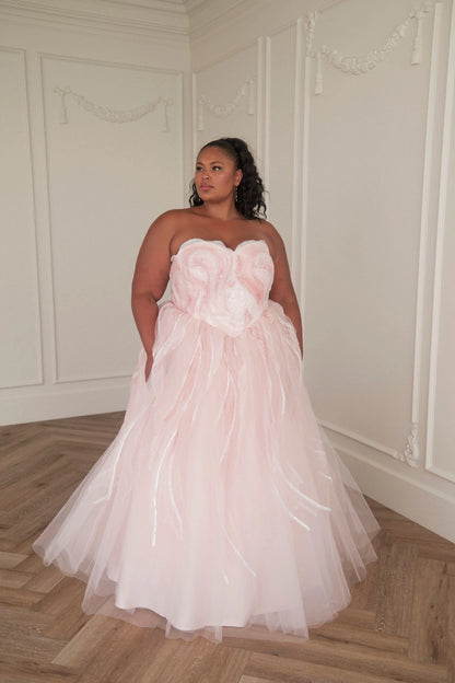 The image shows a woman in a white wedding dress standing in front of a white wall. She is wearing a long, flowing dress with a sweetheart neckline and a full skirt that cascades down to the floor.