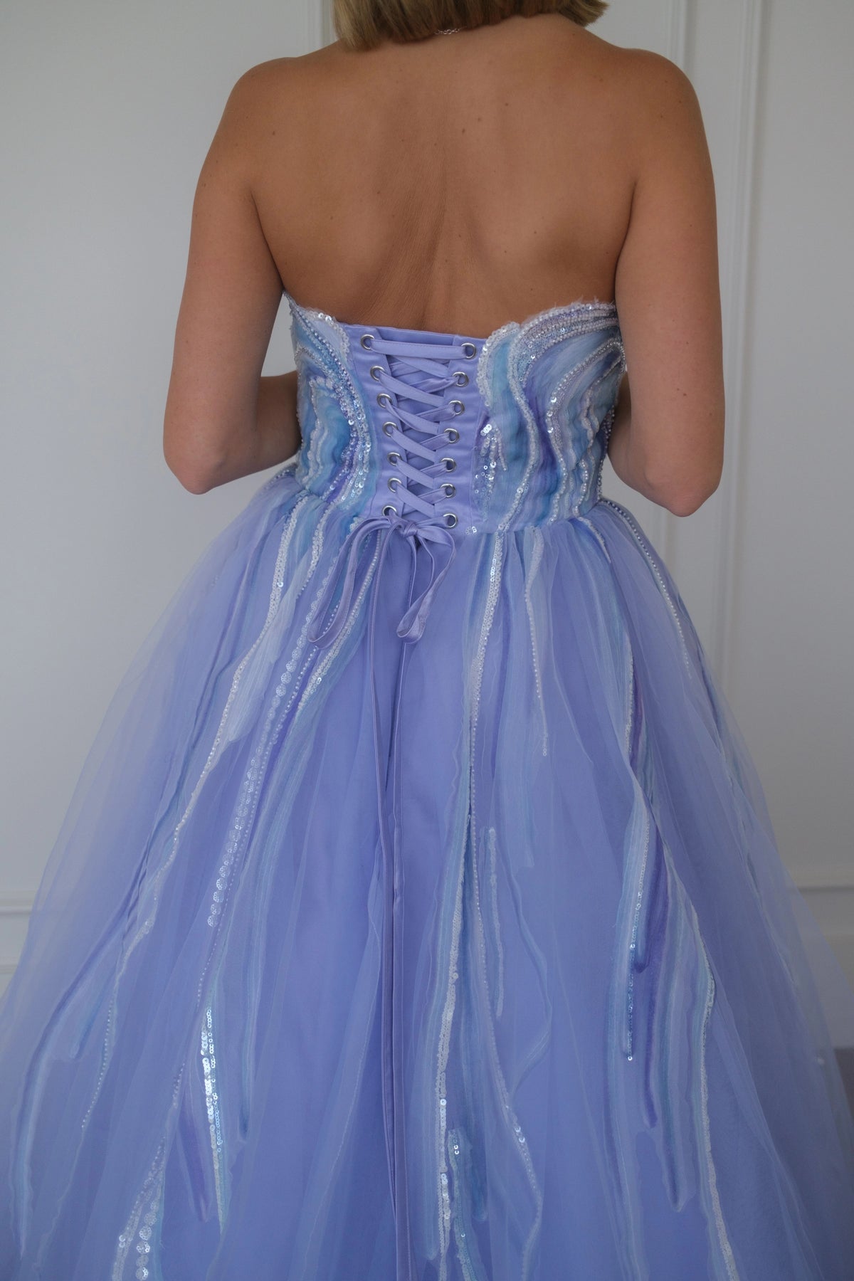 Woman wears a strapless lilac ballgown with corset back and tulle skirt with shiny stripes, facing away.