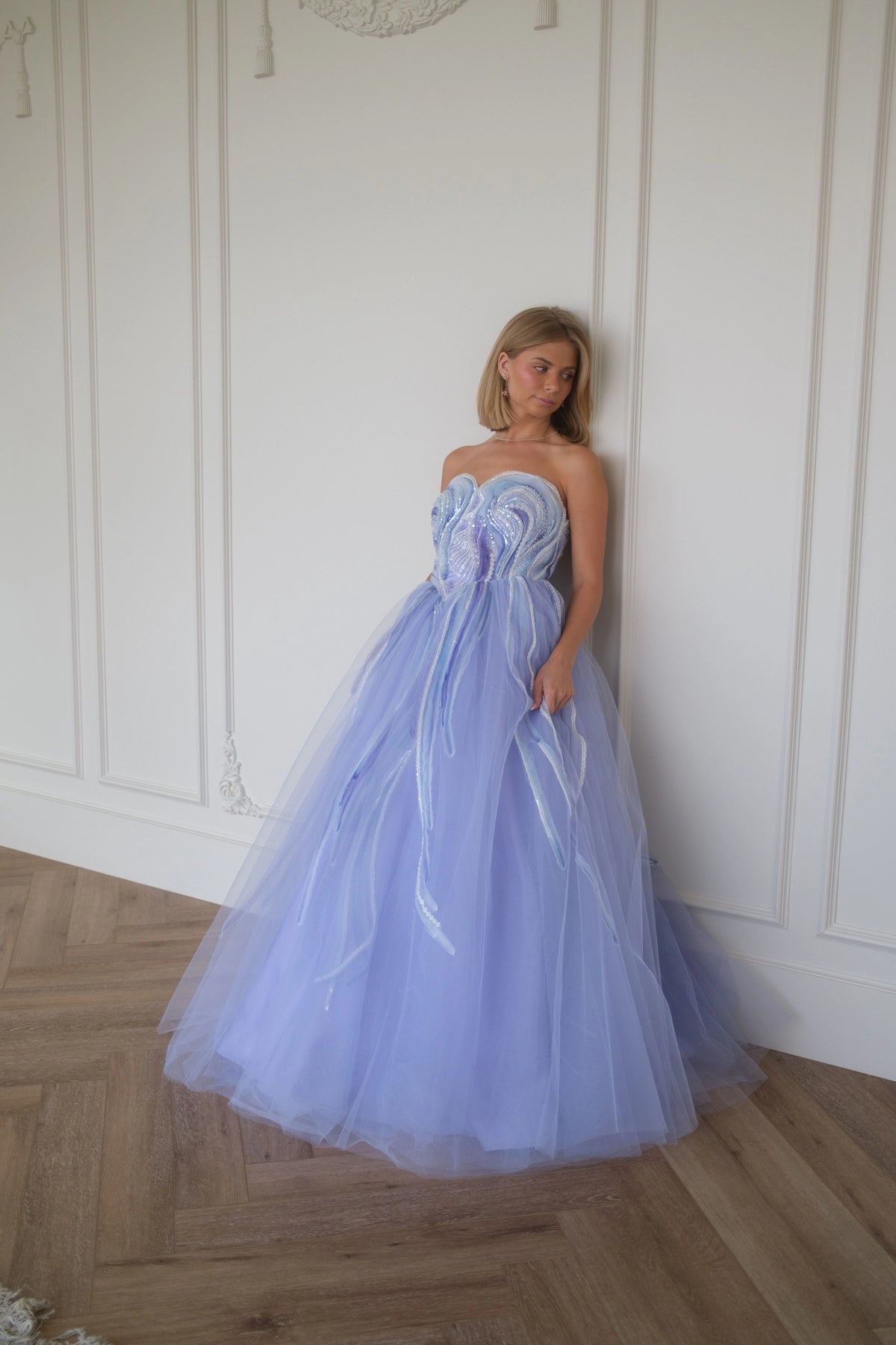 Woman in strapless lavender formal gown with tulle skirt and ruffled bodice leans against white wall on wooden floor.