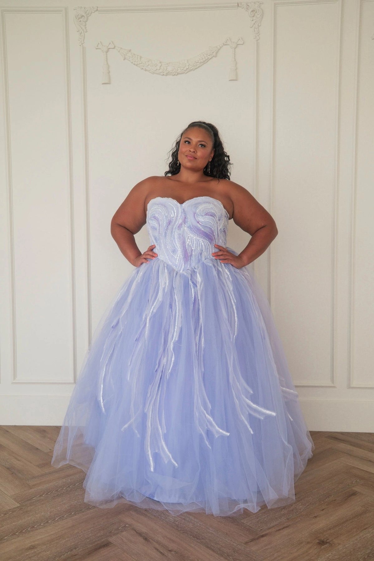 The image shows a woman in a beautiful blue wedding dress standing in front of a white wall, with a floor at the bottom. She is wearing a stunning dress with intricate details and a long train, and