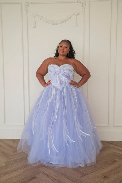 The image shows a woman in a beautiful blue wedding dress standing in front of a white wall, with a floor at the bottom. She is wearing a stunning dress with intricate details and a long train, and